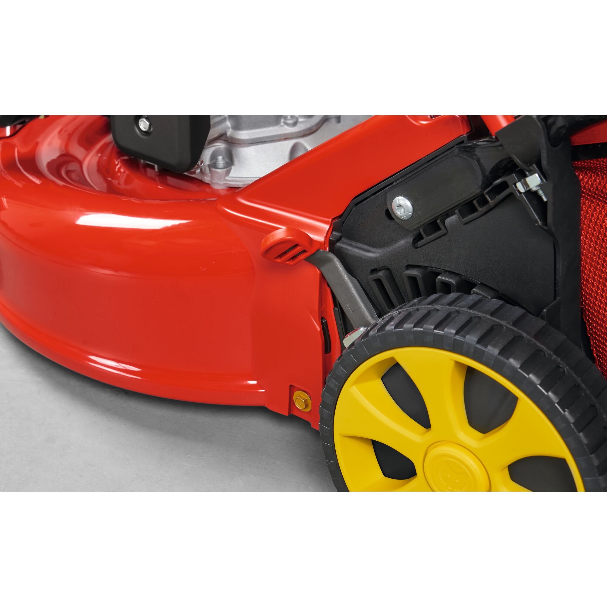 Close-up view of a lawn mower with a red body and a yellow wheel on a concrete surface.