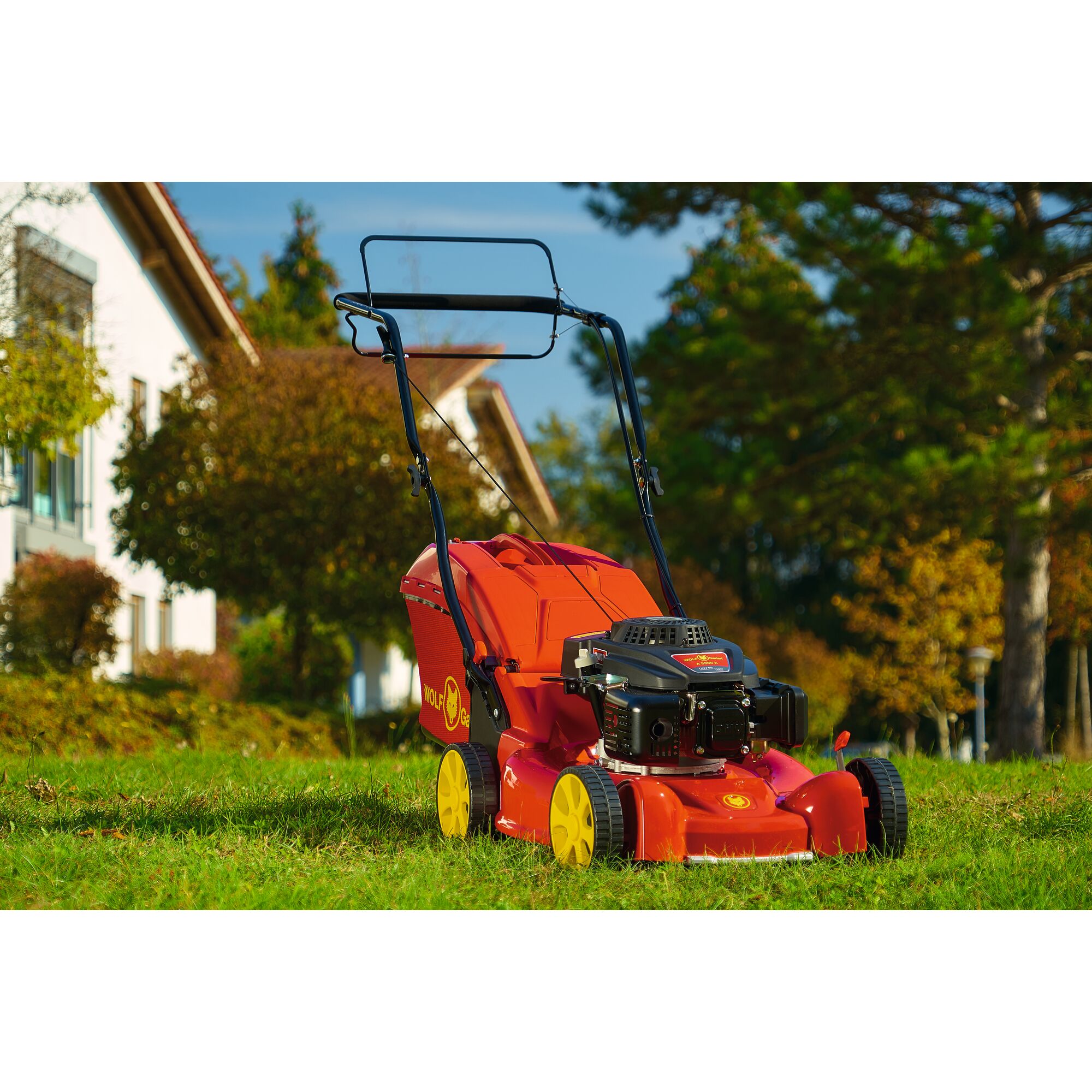 A red and yellow WOLFGarten lawn mower with a grass collection box standing on a grassy lawn outdoors, with trees and a house in the background.