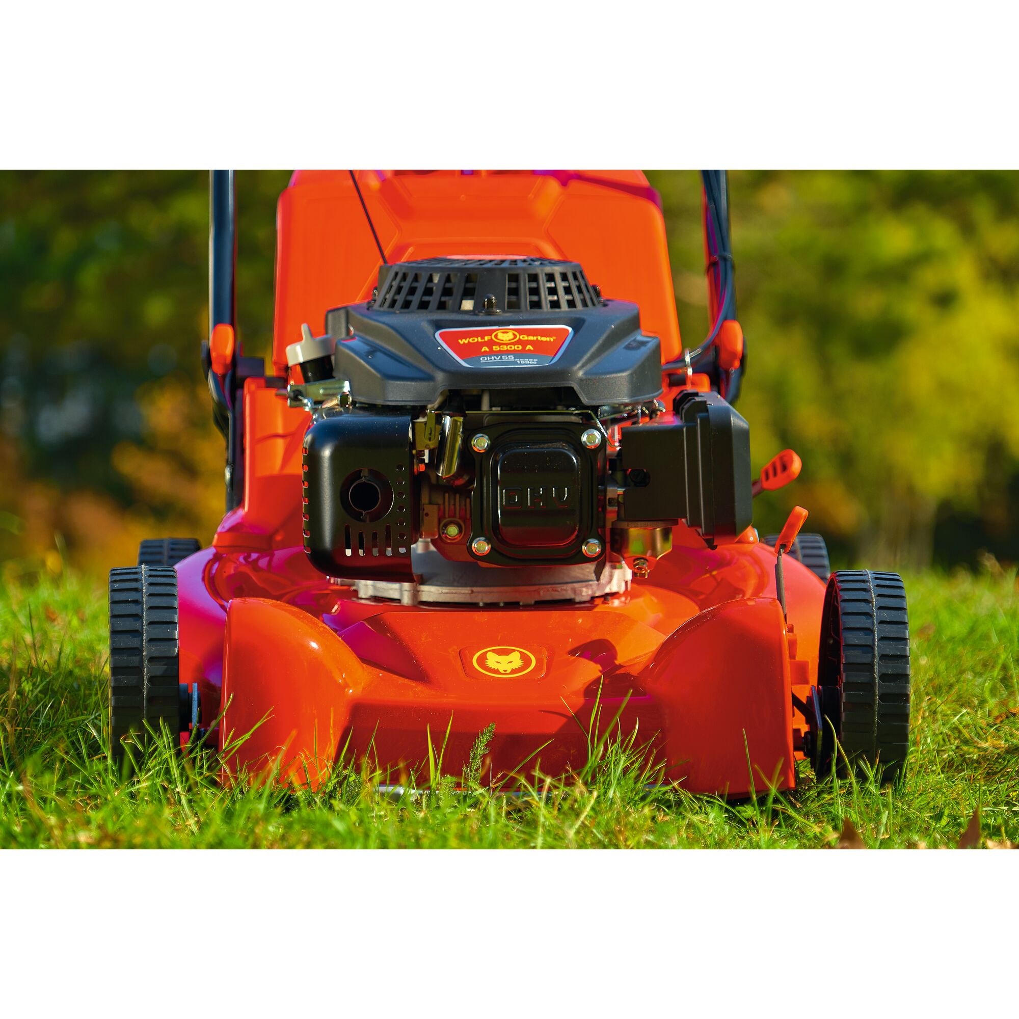 Close-up front view of a WOLFGarten lawn mower with a bright red body, black engine, and visible wheels, positioned on green grass outdoors.