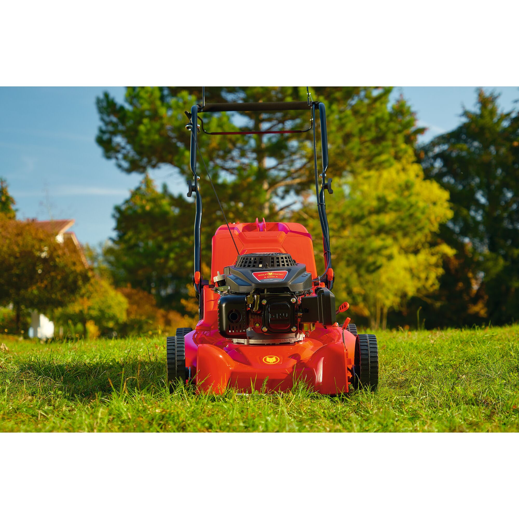 A red lawn mower positioned on green grass outdoors, with trees and a house in the background.