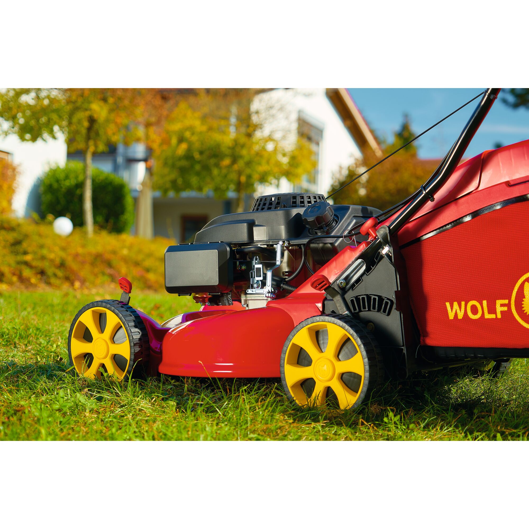 A close-up view of a red WOLFGarten lawn mower with yellow wheels, shown cutting grass in a garden, with a house and autumn trees in the background.
