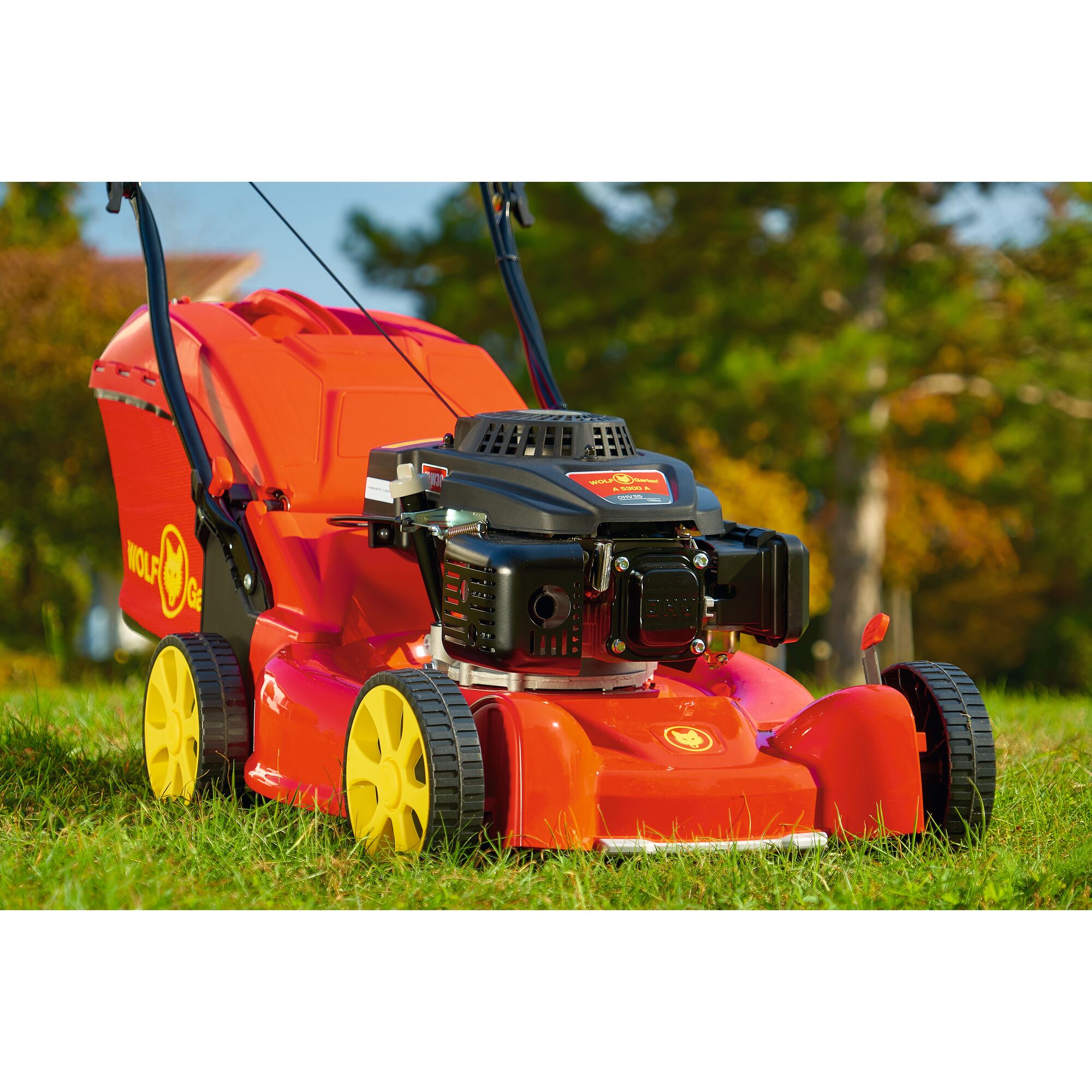 A red and yellow WOLFGarten lawn mower with a grass collection box, photographed outdoors on green grass with trees in the background.