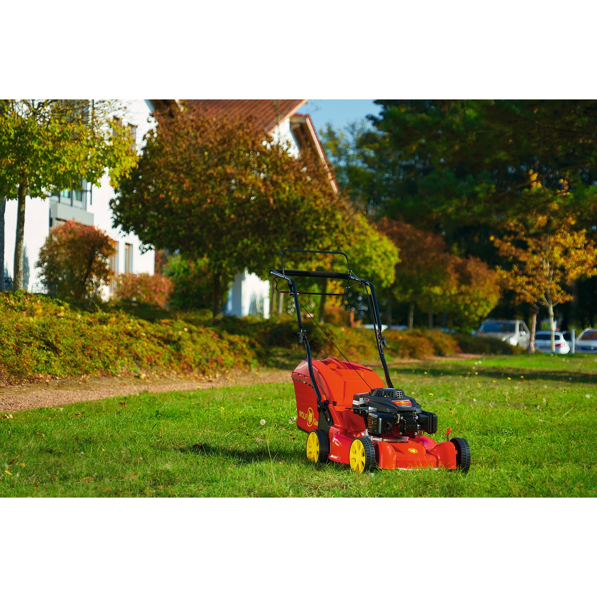 A WOLFGarten lawn mower with yellow wheels and red body stands on green grass in a residential area with trees and houses in the background.