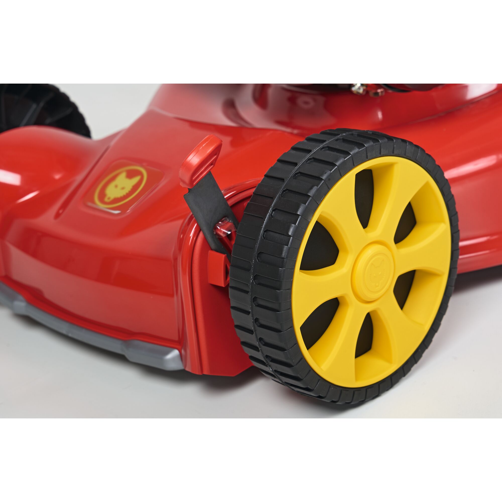 Close-up of the side of a red lawn mower with a yellow wheel and black tread. A lever is attached near the wheel, and a logo with a wolf head is visible on the mower body.