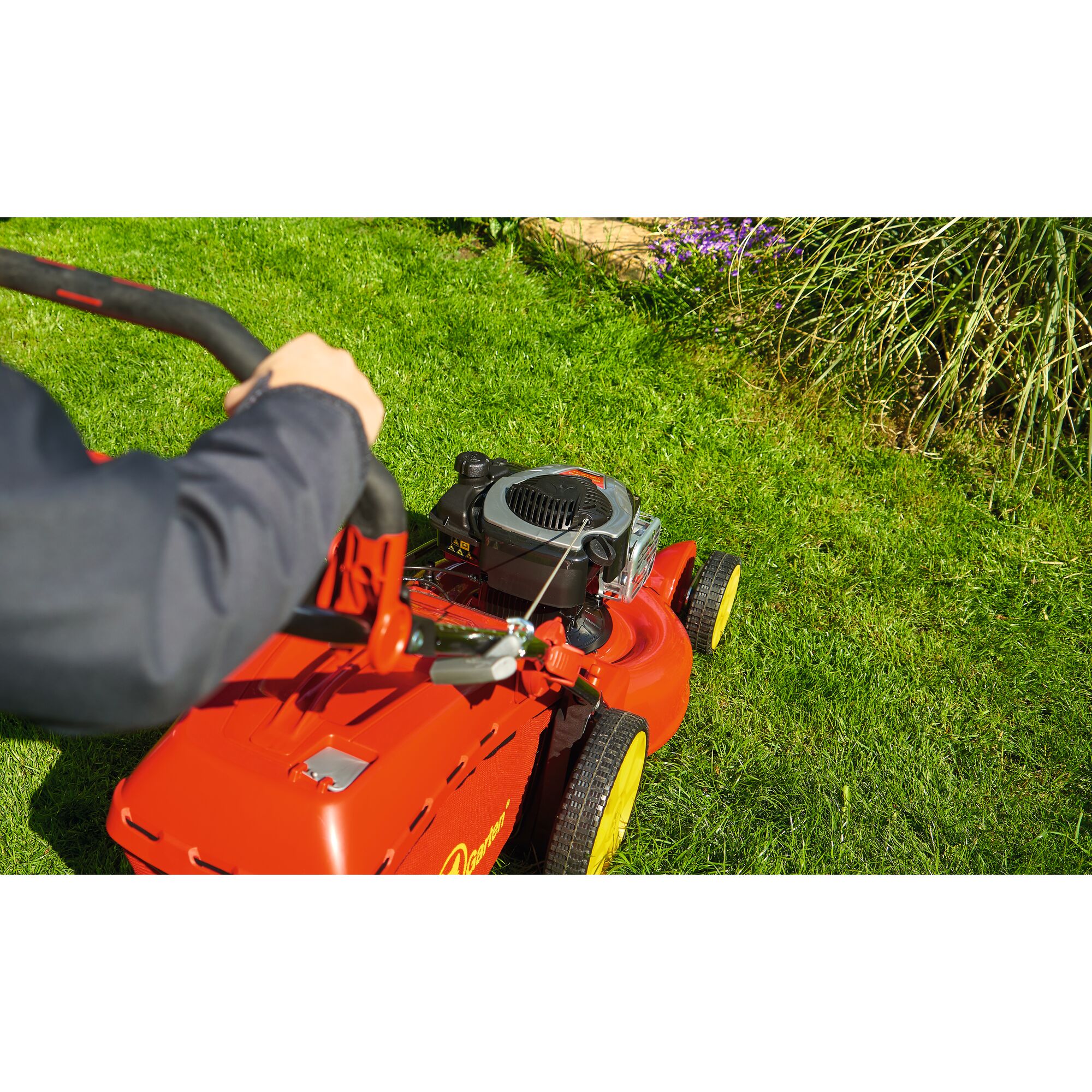 A person is pushing a red WOLFGarten lawn mower with yellow wheels on green grass in a garden.
