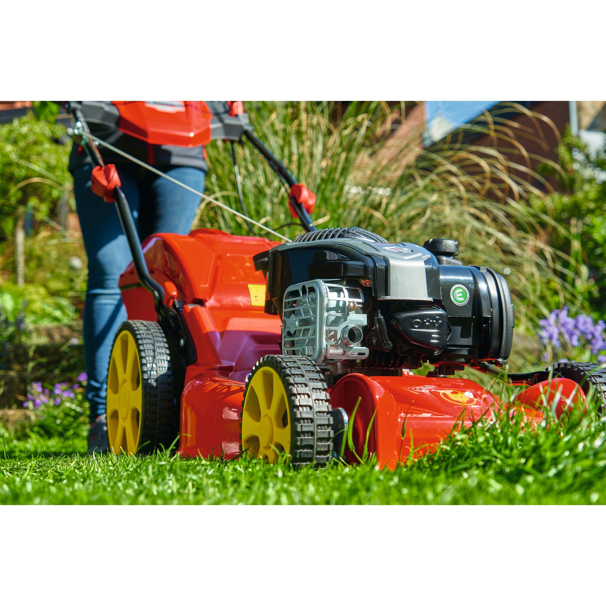 A close-up of a red and yellow lawn mower with large wheels, being pushed on green grass in a garden. The operator's legs are visible in the background.