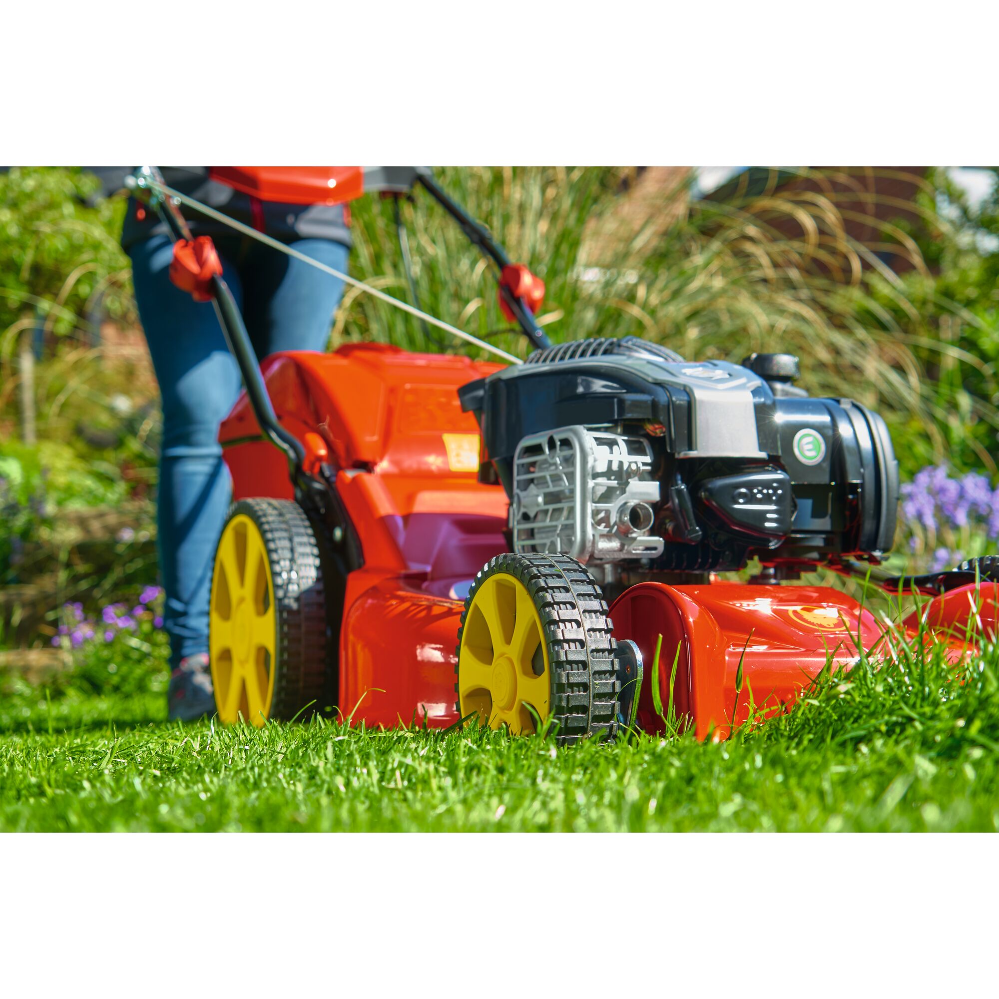 Close-up view of a person using a bright red lawn mower with yellow wheels to cut grass in a garden.