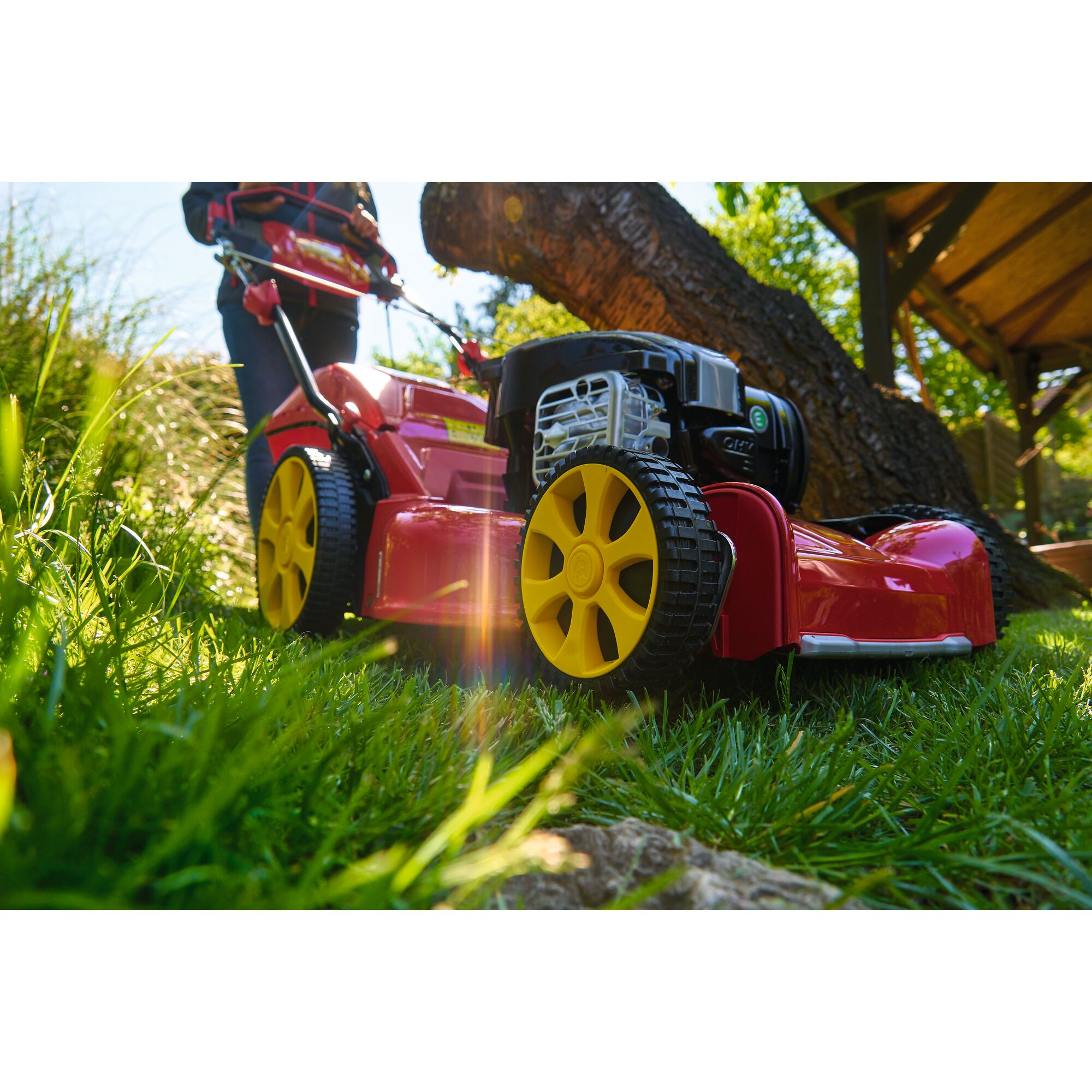 Close-up view of a red and yellow lawn mower on green grass, with a person standing behind it outdoors.