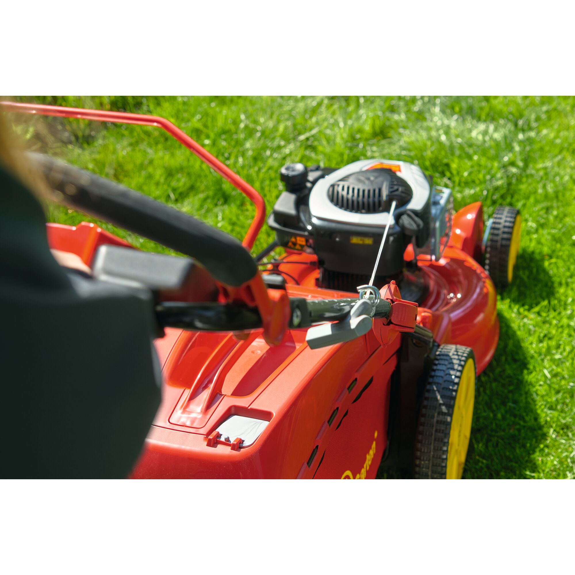 Close-up view of a red and yellow lawn mower on green grass, showing the handle, control lever, and part of the engine.