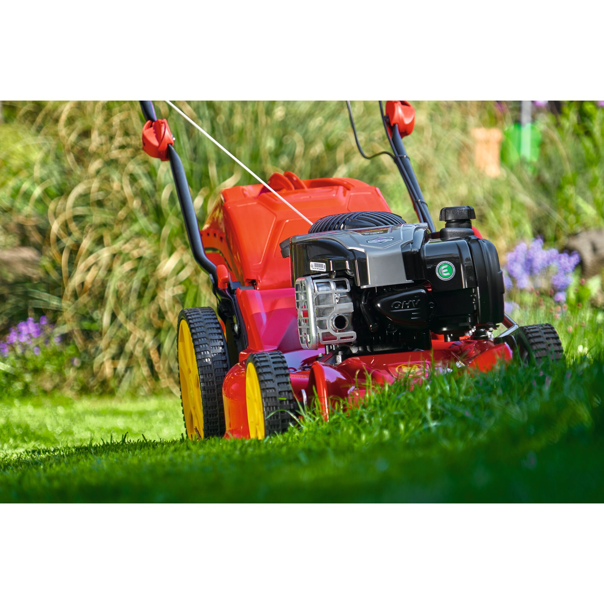 Close-up view of a red and yellow lawn mower with a black engine, positioned on green grass in a garden with plants and flowers in the background.