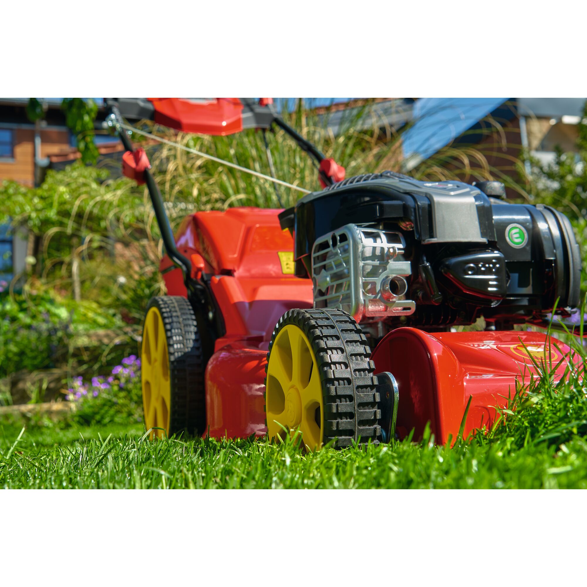Close-up view of a red and yellow lawn mower on green grass in a garden, with the house and plants visible in the background.