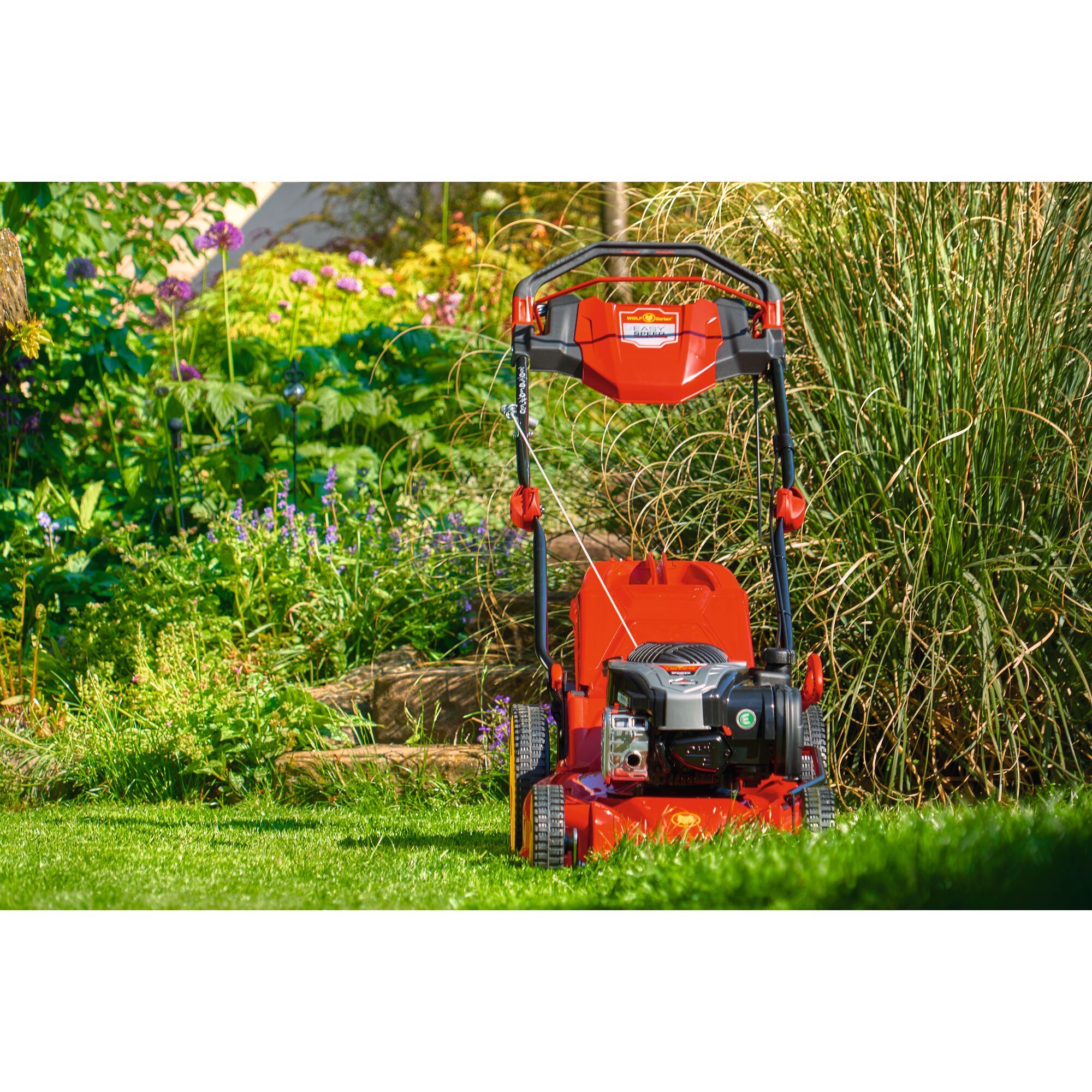 A red WOLFGarten lawn mower stands on a grassy lawn in a garden with tall plants and flowers in the background.