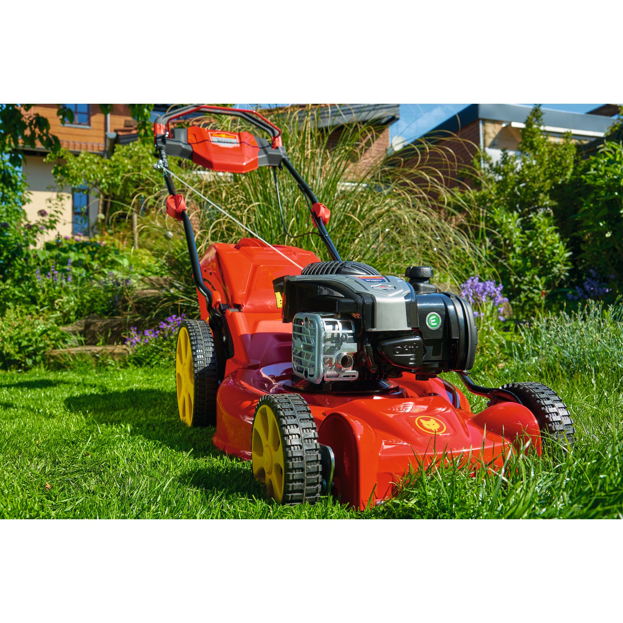 A red and yellow lawn mower is positioned on a green lawn in a garden, with plants and a house visible in the background.