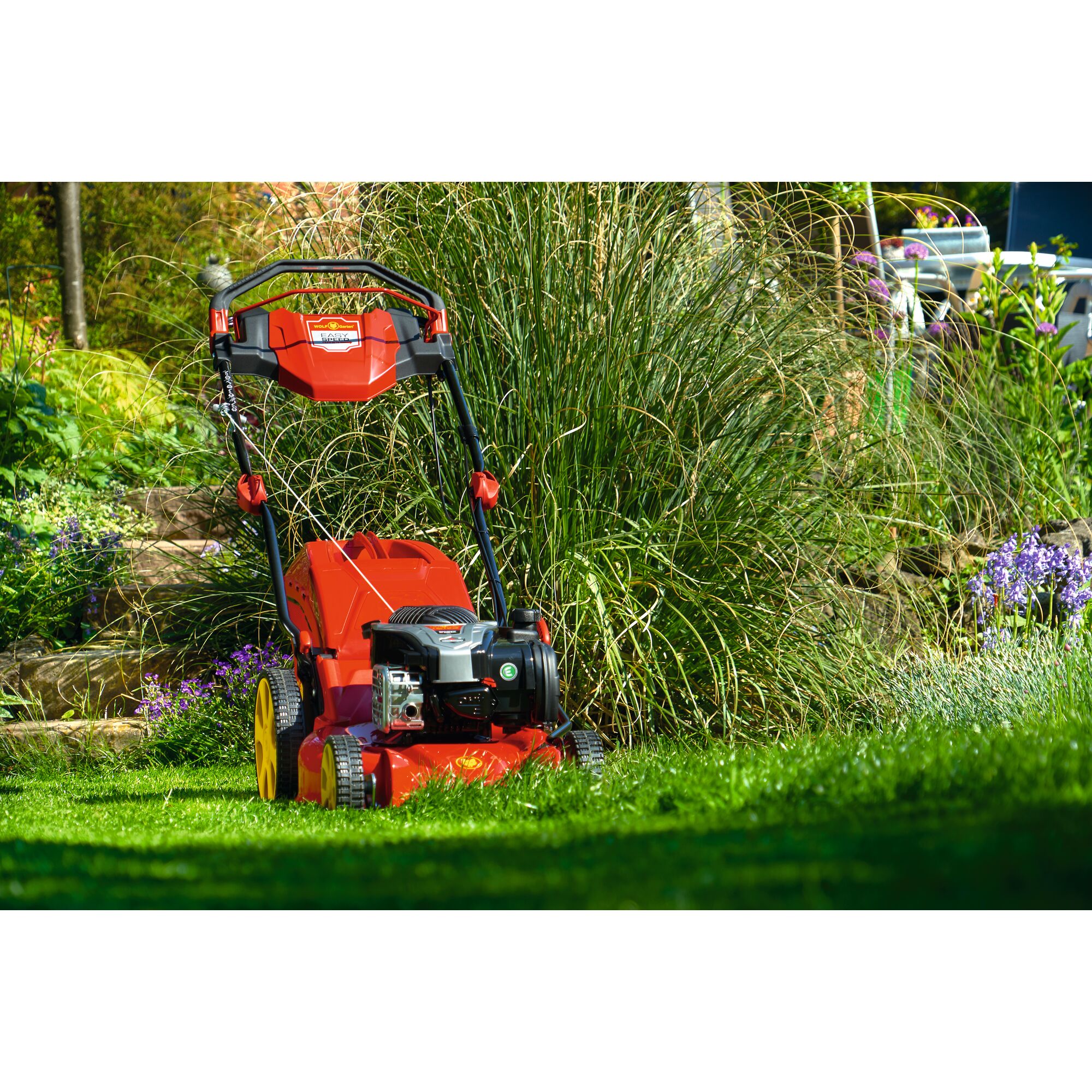 A red and yellow WOLFGarten lawn mower is placed on green grass in a garden surrounded by tall plants and flowers.