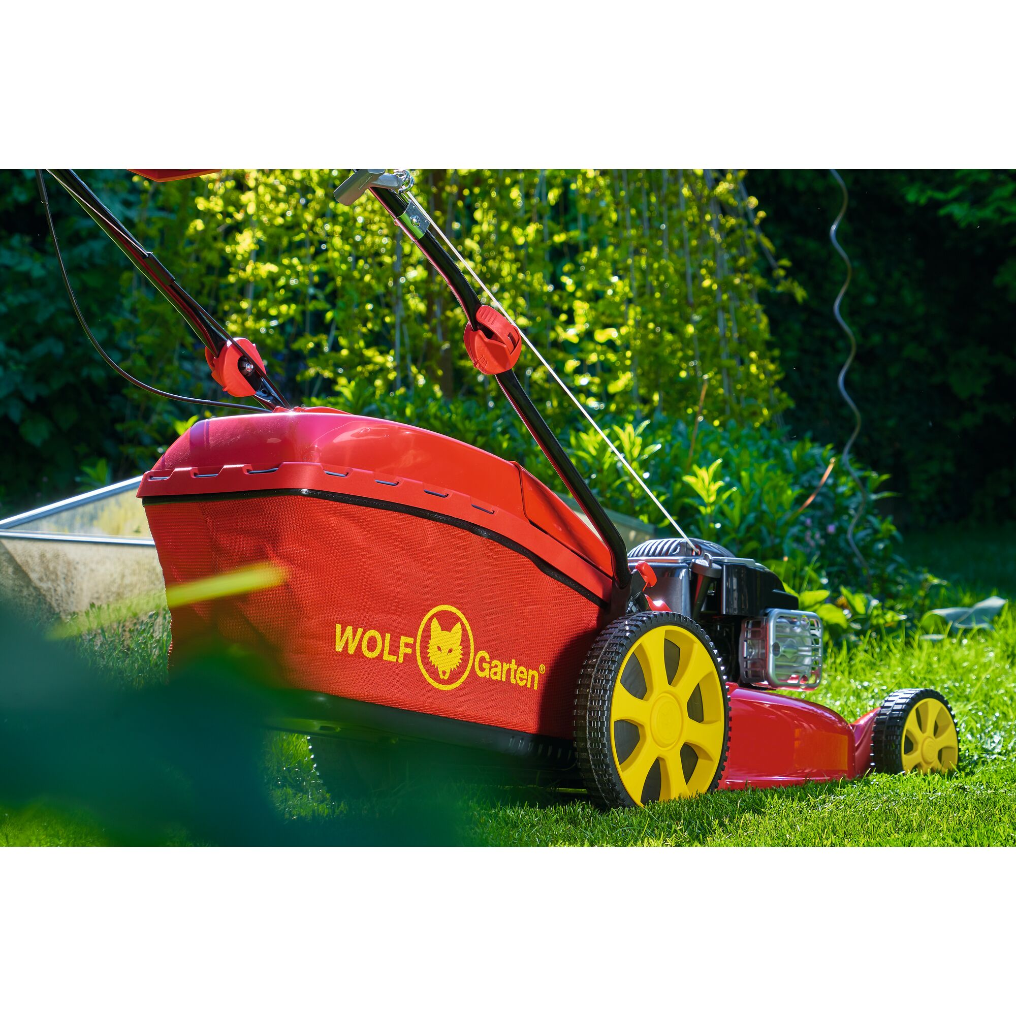 A close-up view of a red and yellow WOLFGarten lawn mower on grass in a garden. The lawn mower features prominent yellow wheels and a grass collection bag with the WOLFGarten logo printed in yellow.