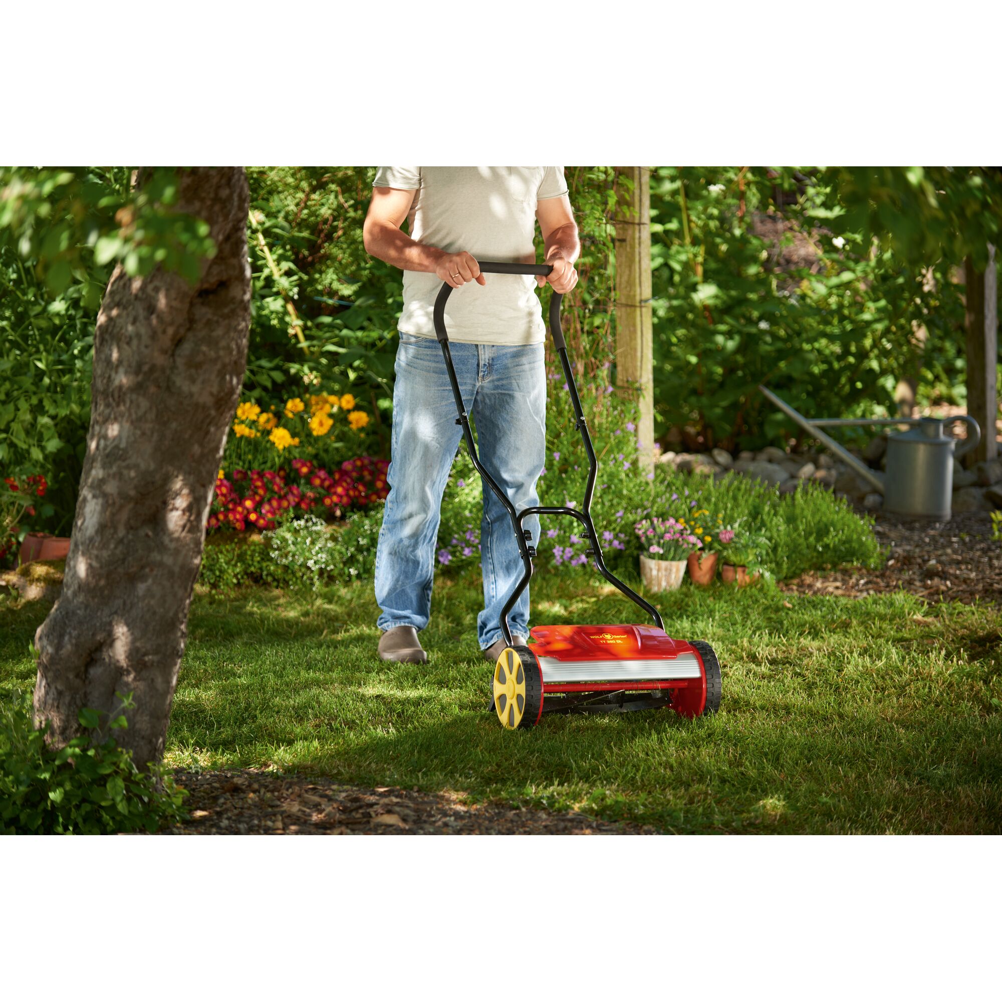 A person uses a red and yellow manual lawn mower on a grassy lawn in a garden, surrounded by colorful flowers and greenery.