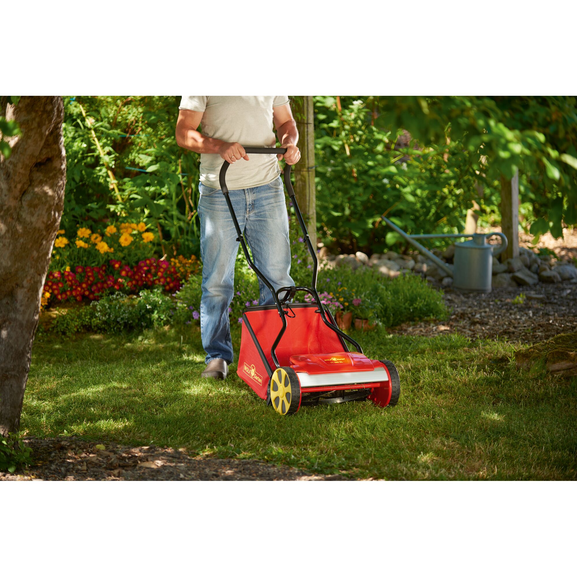 A person using a red and yellow manual lawn mower on a grassy area in a garden with flowers and greenery.
