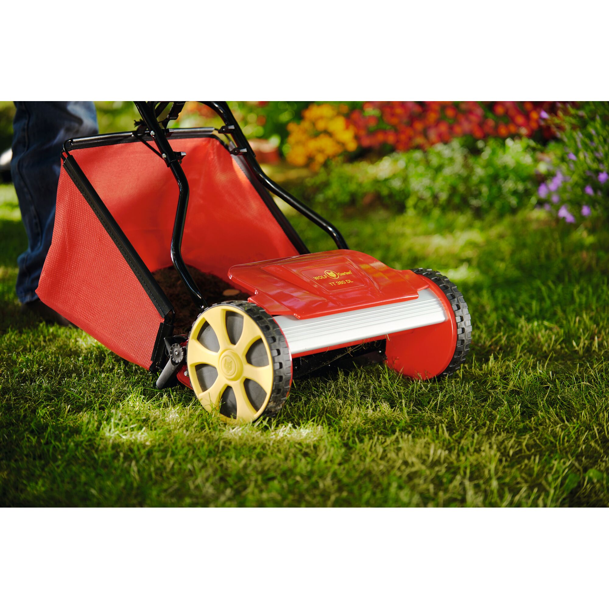 A close-up view of a red and yellow manual lawn mower on green grass, with a person’s legs partially visible and colorful flowers in the background.