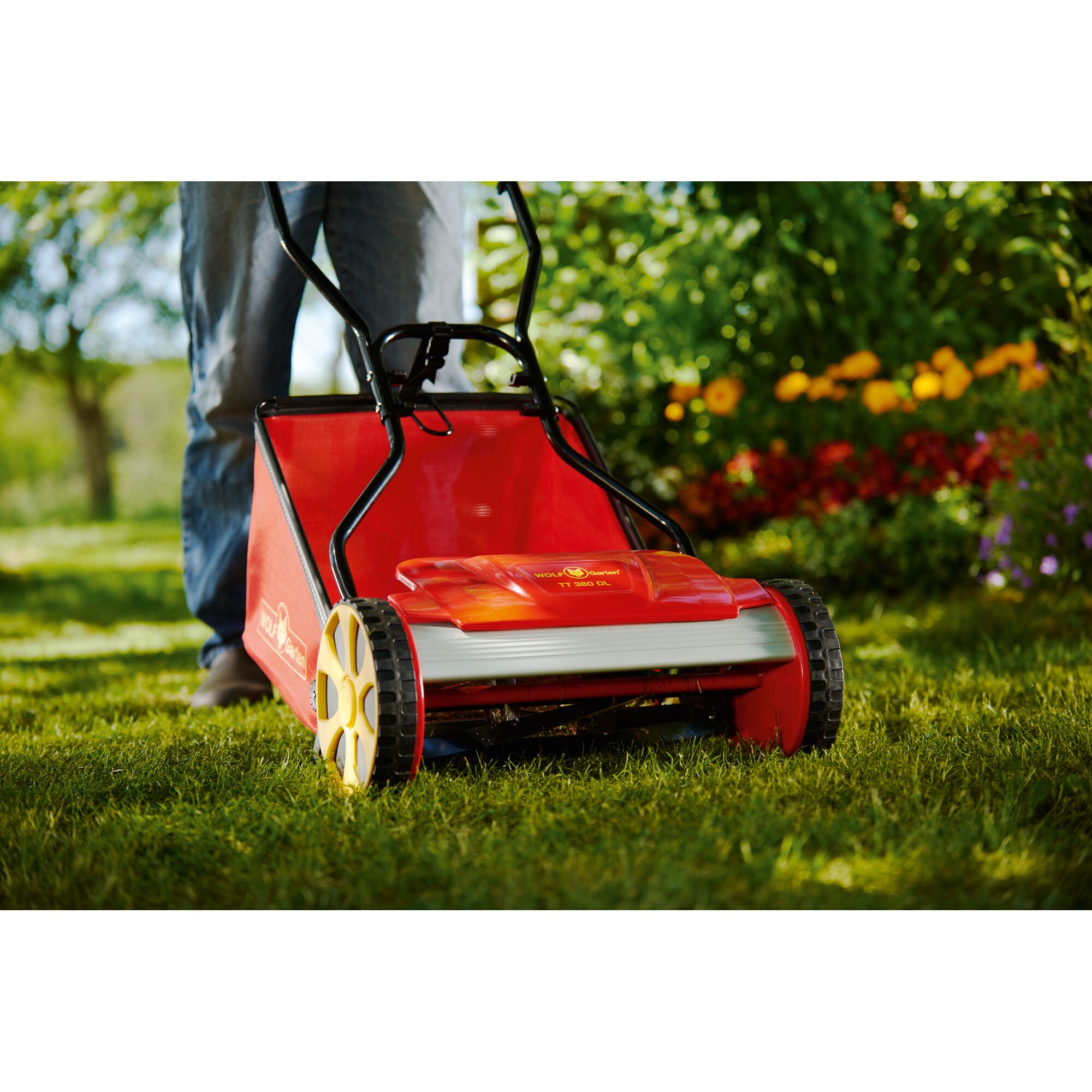 A close-up view of a WOLFGarten manual lawn mower with yellow wheels being used on a green lawn. The mower is bright red and the user’s legs are visible behind it. Colorful flowers and greenery are visible in the background.