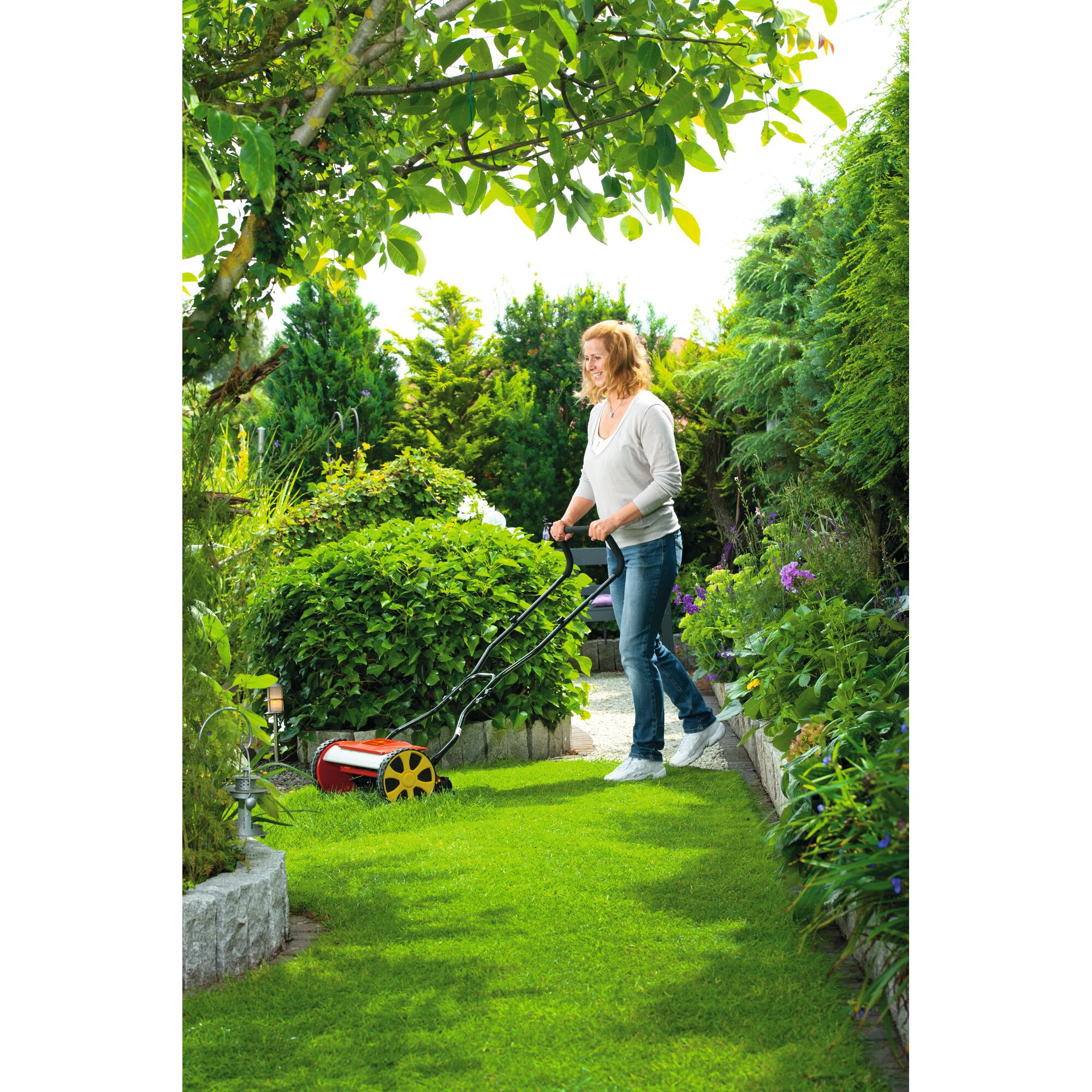 Person pushing a WOLFGarten manual lawn mower with yellow wheels on a grassy lawn surrounded by plants and trees.