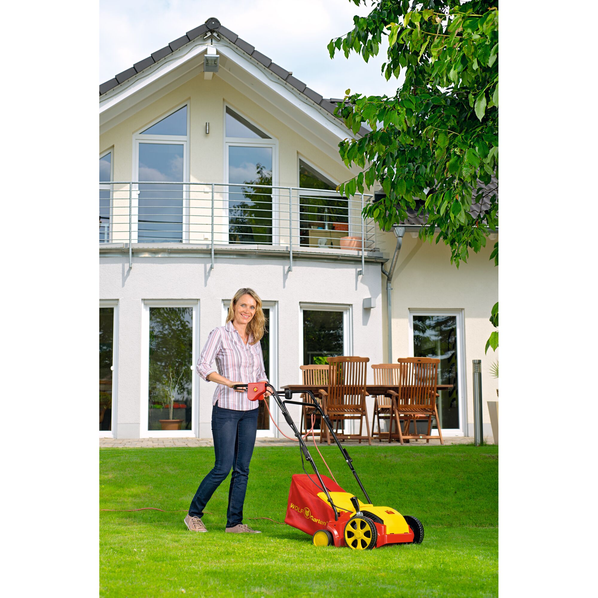 A person is using a WOLFGarten lawn scarifier on a grassy lawn in front of a modern house with large windows and a wooden outdoor dining set.