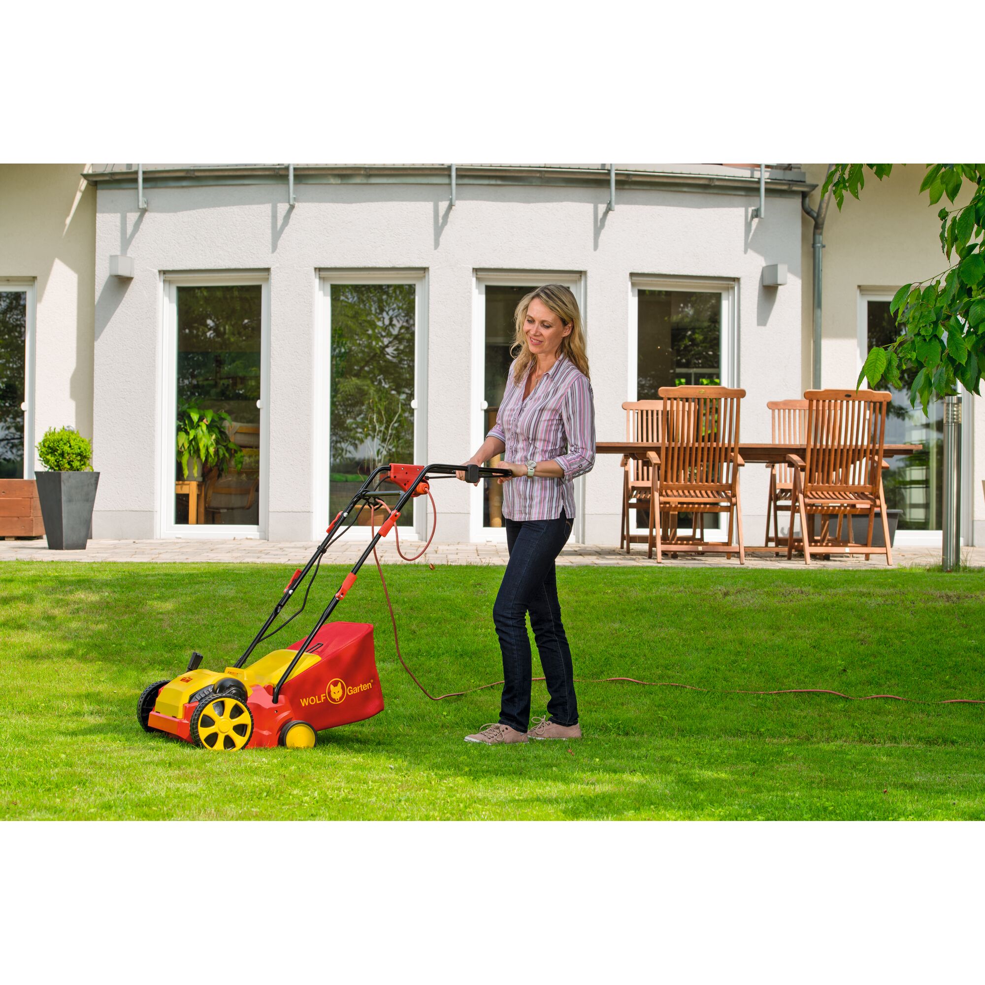 A person is operating a red and yellow WOLF-Garten lawn scarifier on a green lawn in front of a modern house with large windows and wooden garden furniture.