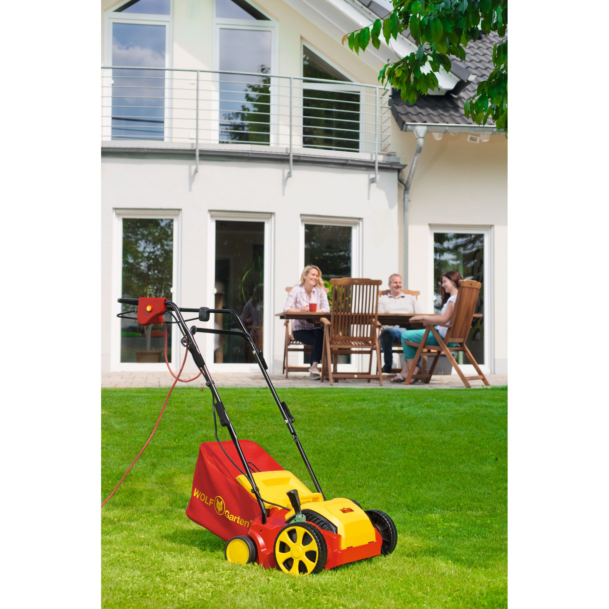 WOLFGarten electric lawn mower with yellow and red design, placed on a green lawn in front of a modern house; three people are sitting at a wooden table on the patio in the background.