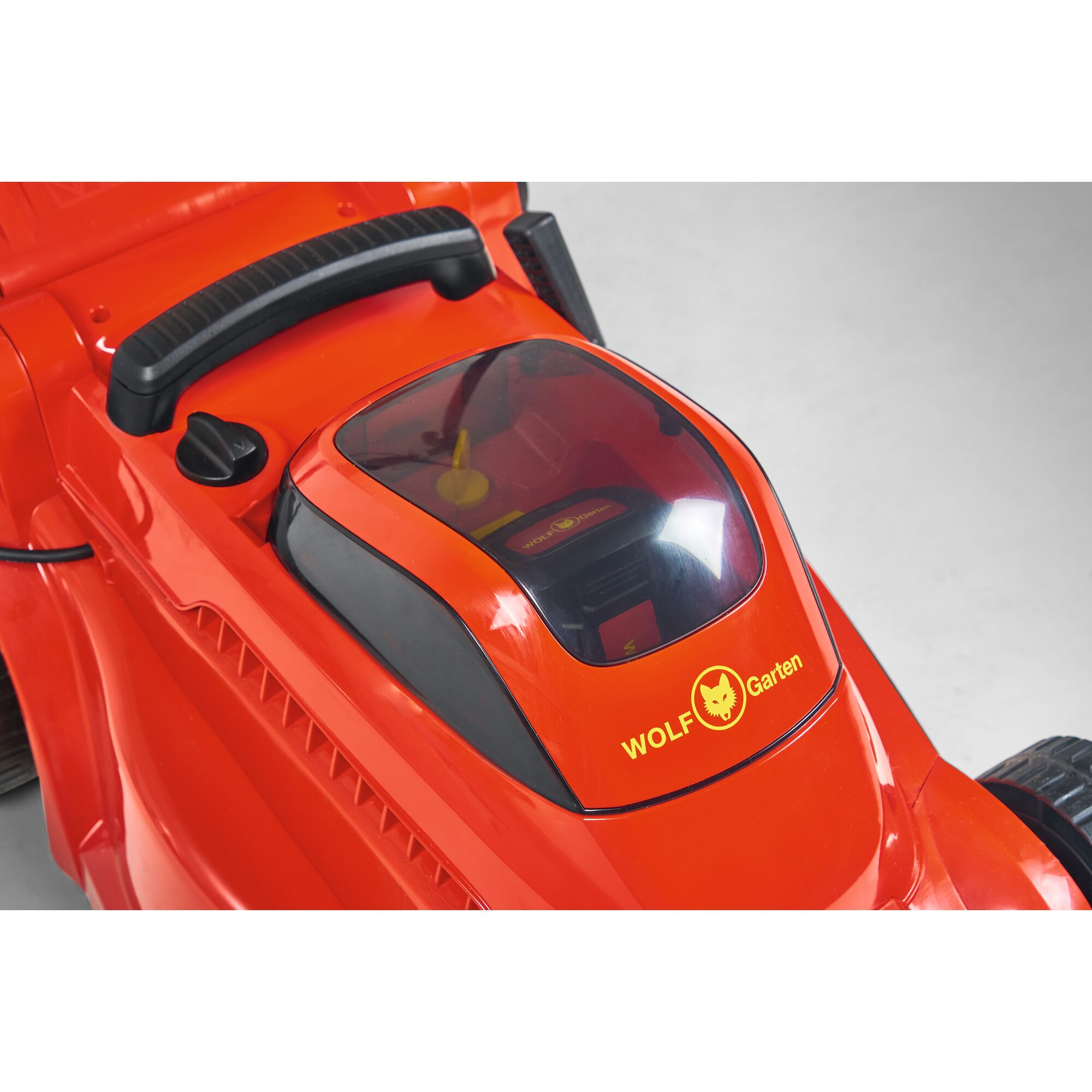 Close-up of a red WOLF Garten lawn mower, focusing on the battery compartment under a transparent cover, with brand logo and handle visible.