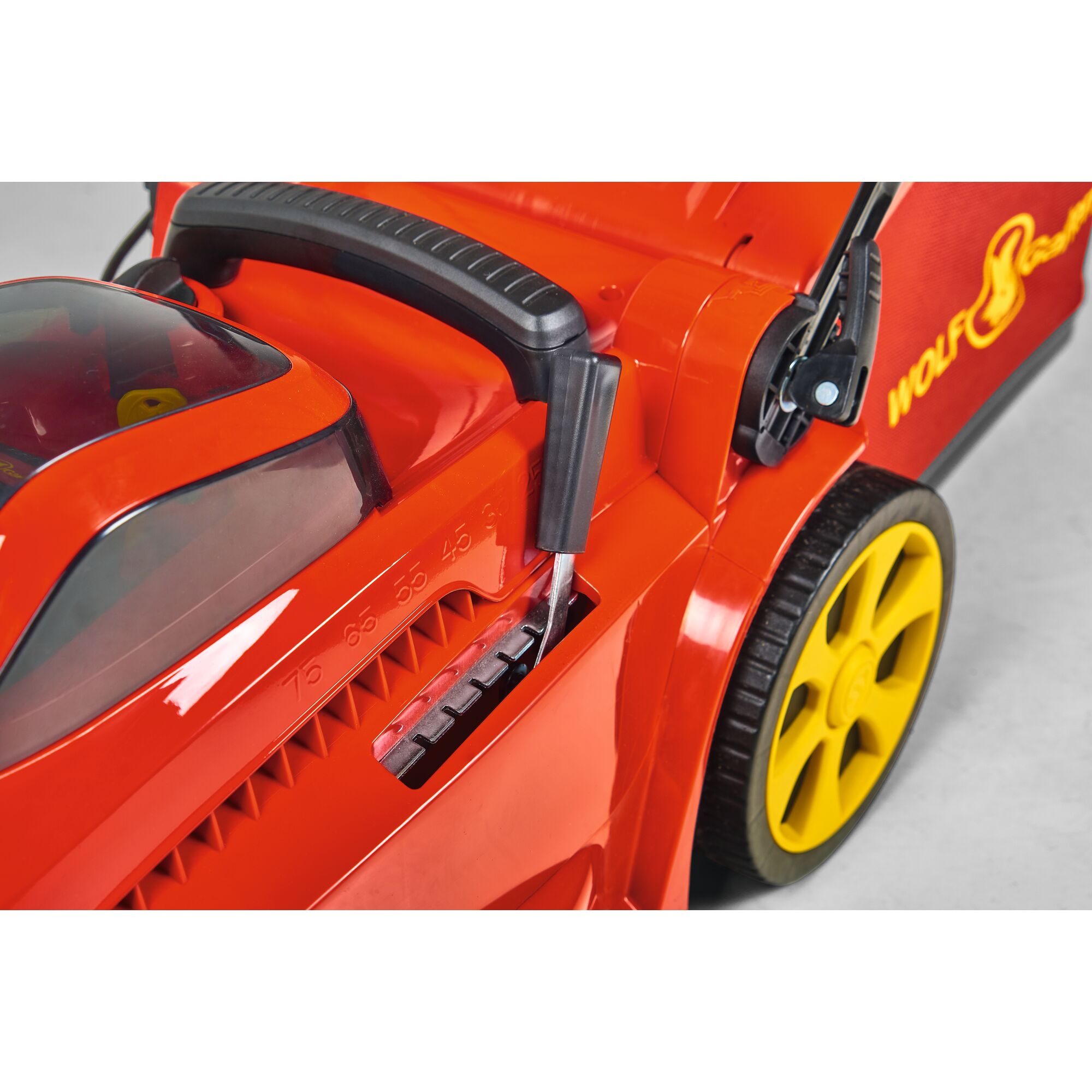 Close-up of a red and yellow WOLFGarten lawn mower, showing the height adjustment lever and part of the rear wheel.