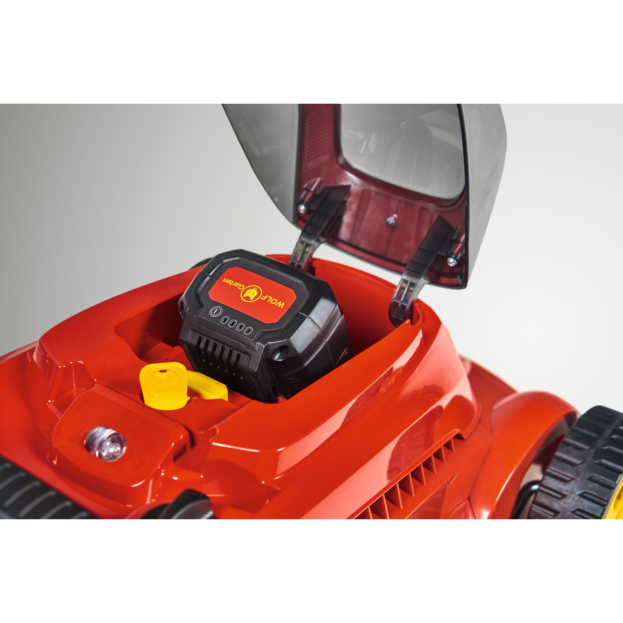 Close-up of a red WOLFGarten lawn mower showing the battery compartment with a WOLFGarten branded battery installed and an open protective cover.