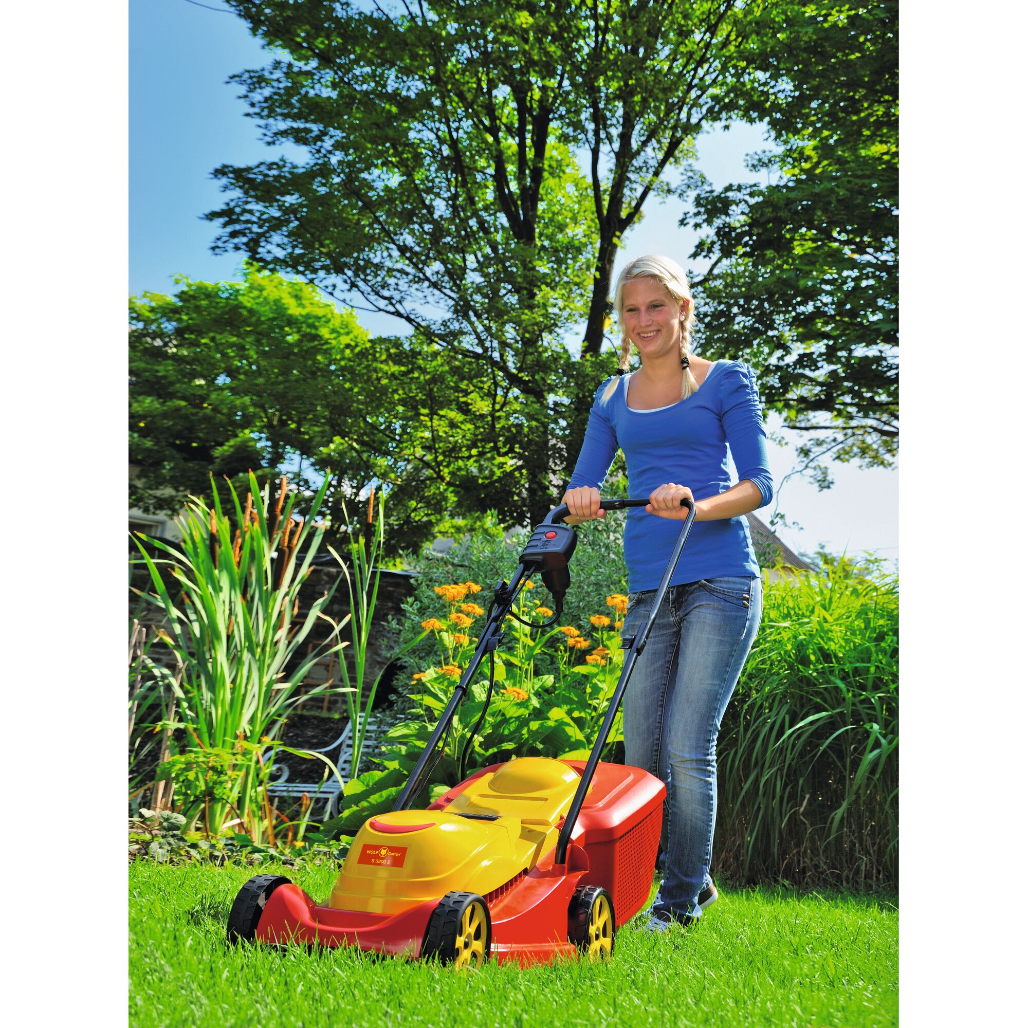 A person with an obscured face wearing a blue top and jeans is pushing a yellow and red WOLFGarten lawn mower across a green garden with tall plants and flowers, surrounded by trees.