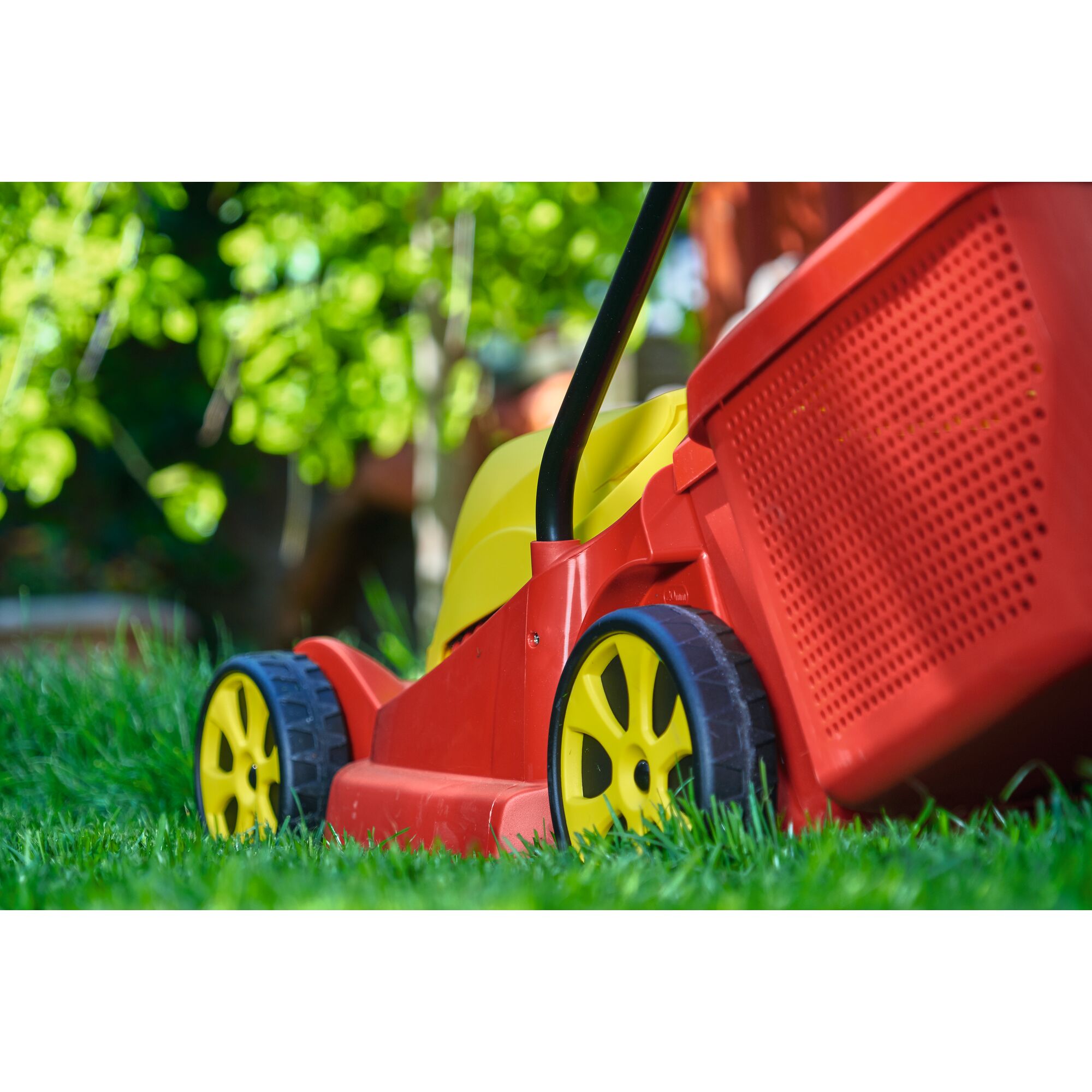 Close-up view of a red and yellow lawn mower with a black handle, positioned on green grass in a garden.