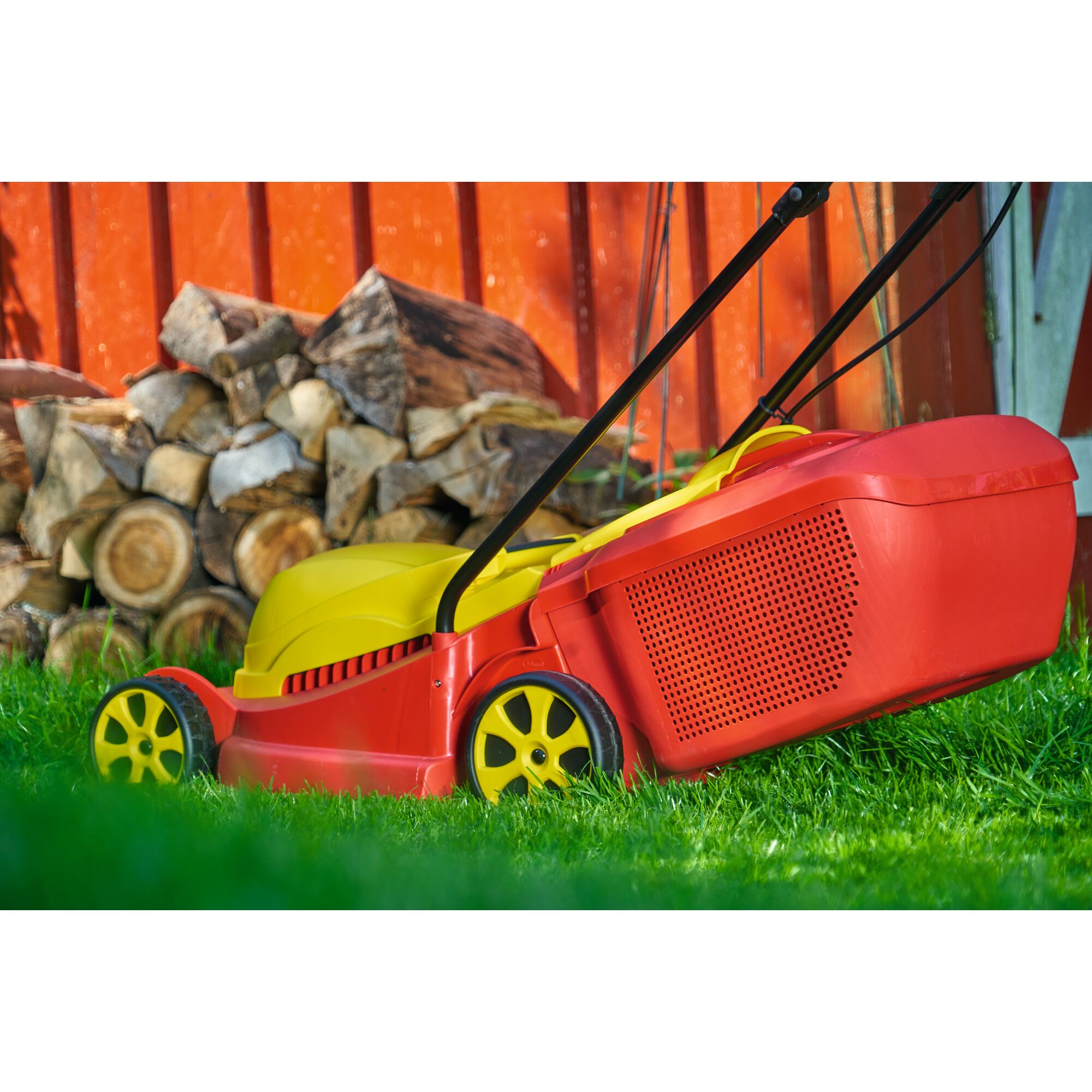 A red and yellow lawn mower on green grass, with a stack of chopped firewood and an orange wooden fence in the background.