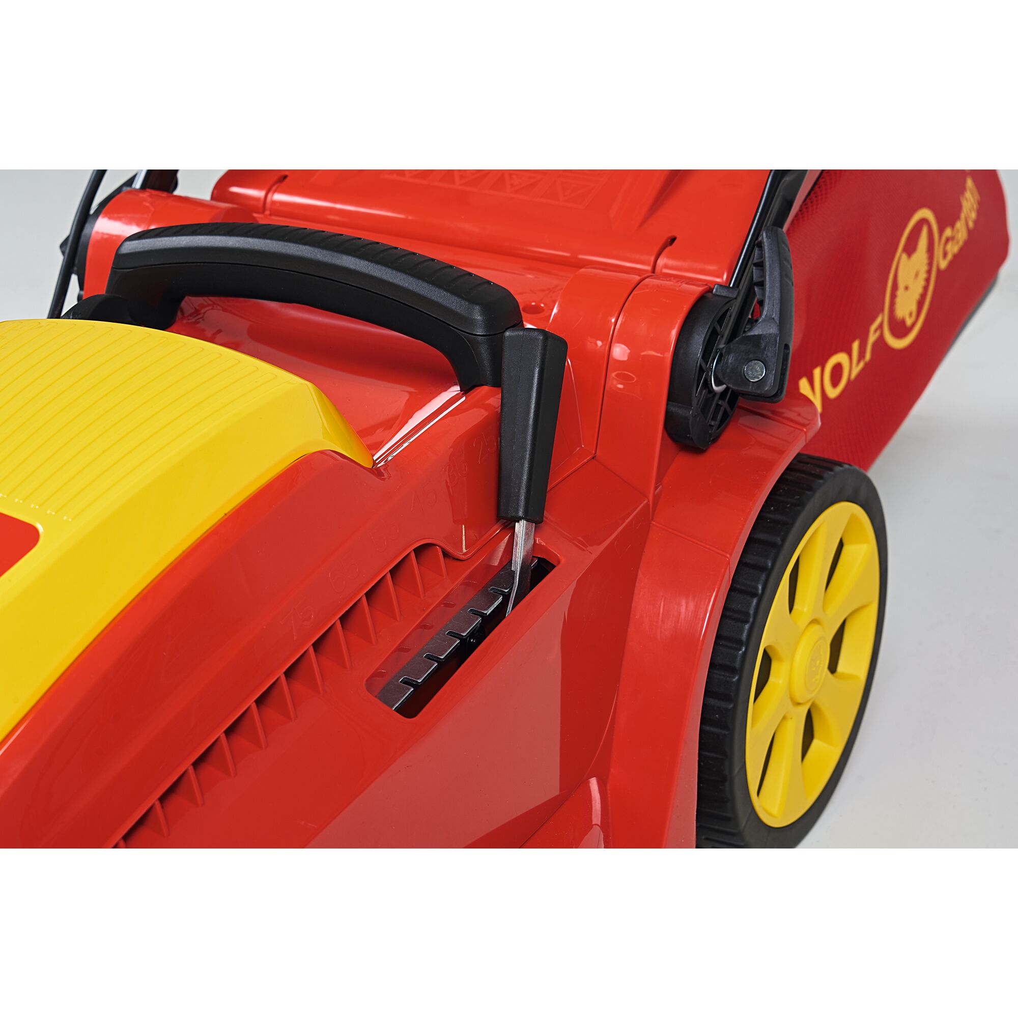 Close-up of a red and yellow WOLFGarten lawn mower, showing the side lever for adjusting cutting height, a black handle, yellow wheel, and part of the grass collection bag with the WOLFGarten logo.
