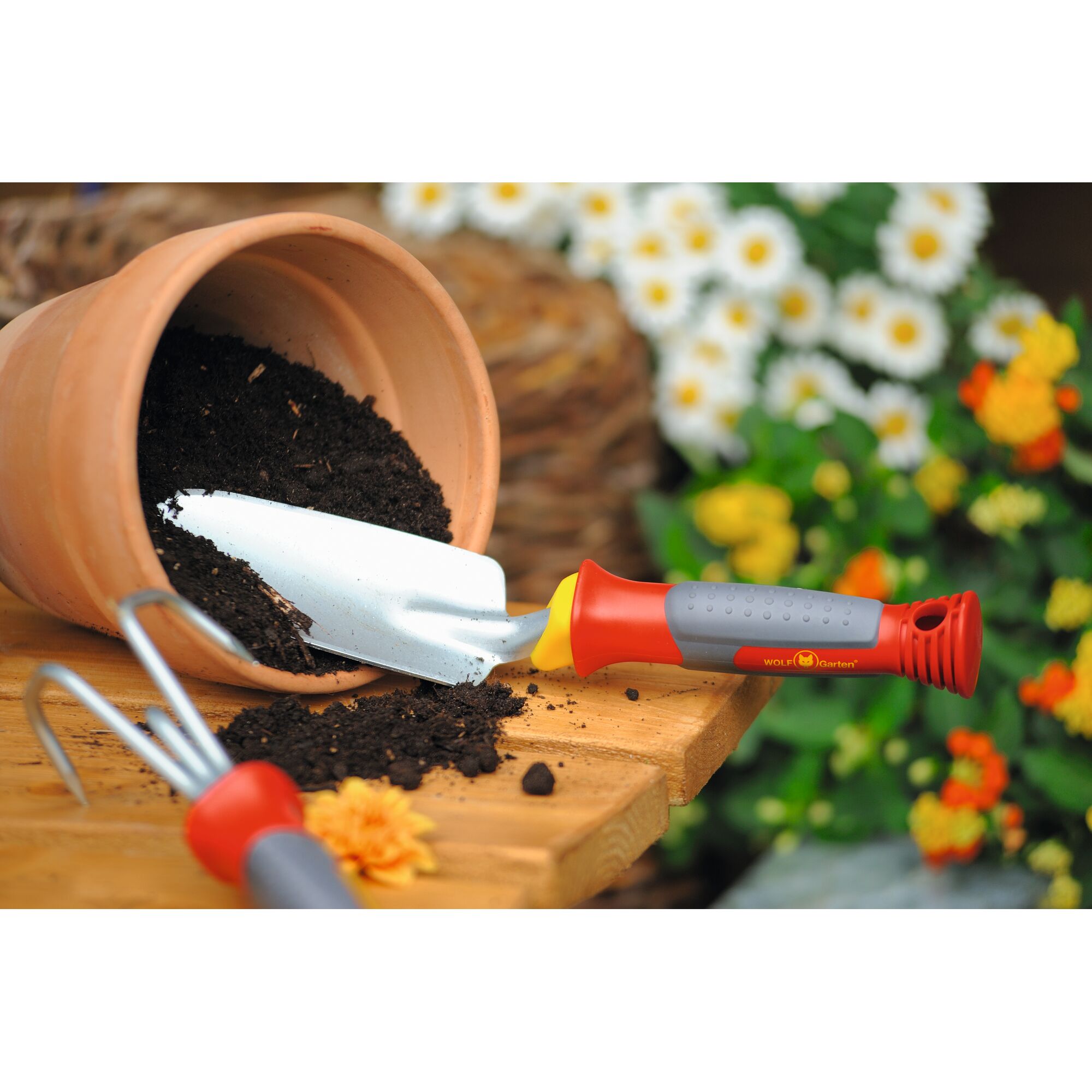 A WOLFGarten gardening hand trowel with a red and grey handle is seen on a wooden surface next to a terracotta pot filled with soil, surrounded by garden flowers.