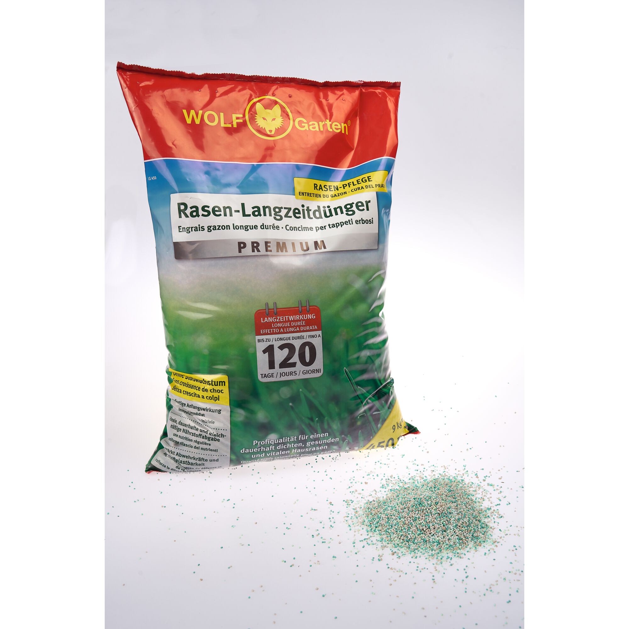 A bag of WOLF Garten Rasen-Langzeitdünger lawn fertilizer shown upright, with scattered fertilizer granules on the white surface in front of it. The packaging highlights the product's premium quality and its long-lasting effect for up to 120 days.