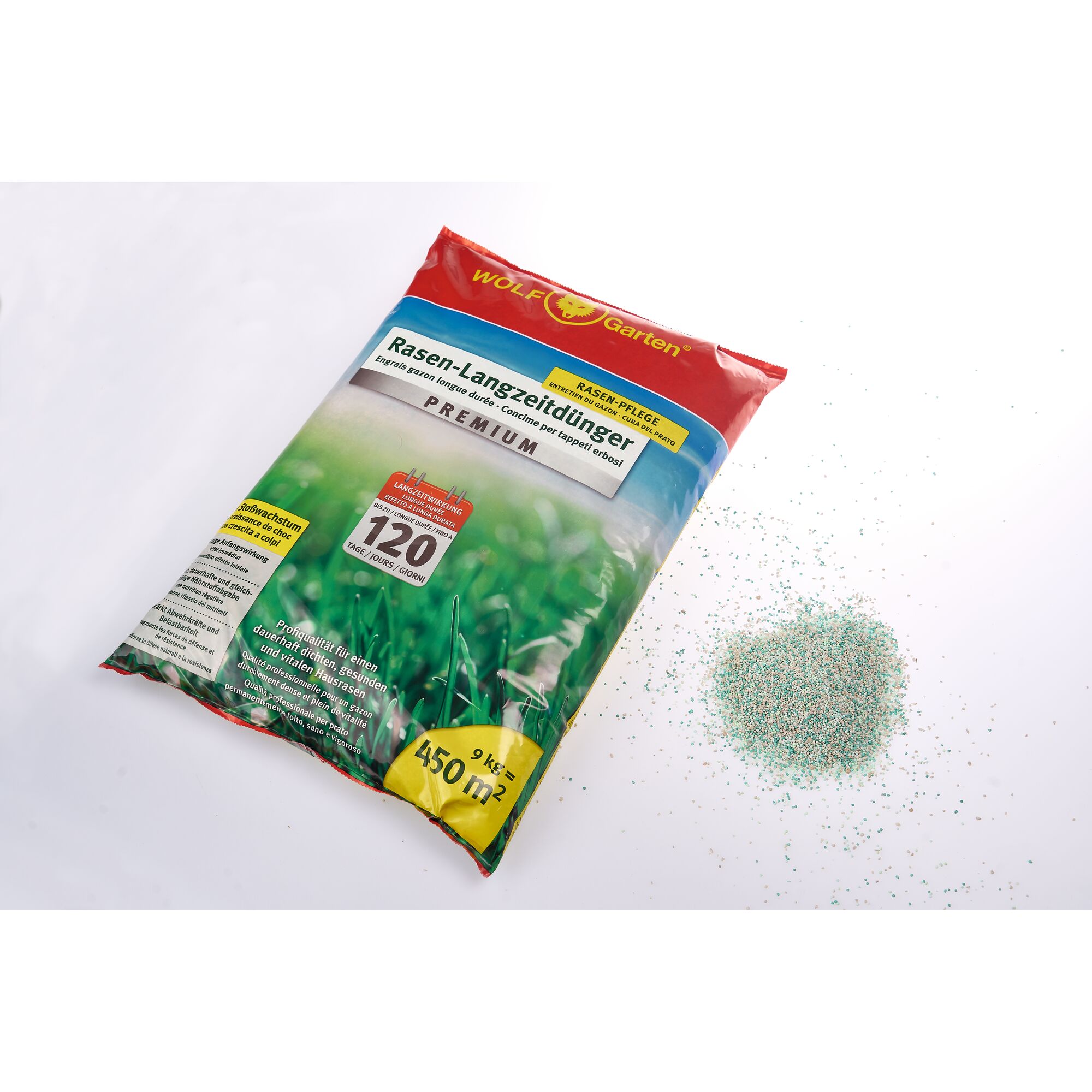 A bag of WOLF Garten Rasen-Langzeitdünger Premium lawn fertilizer is shown on a white surface. The bag displays product information including coverage of 450 m² and 9 kg content. Next to the bag, there is a small pile of multicolored granular fertilizer.
