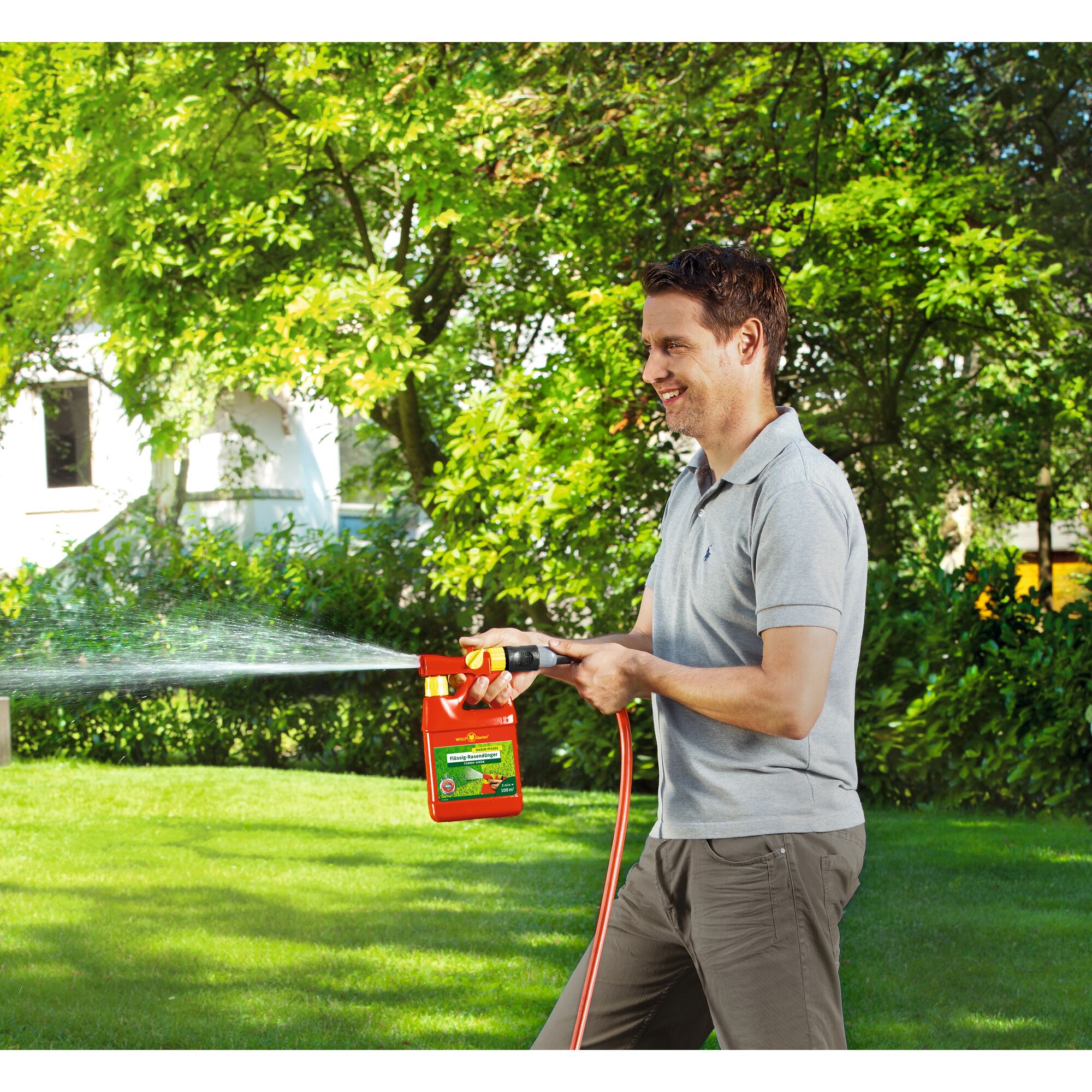 A person with a blurred face is using the WOLFGarten LT100BDA garden sprayer attached to a hose, spraying water onto grass in a green garden.