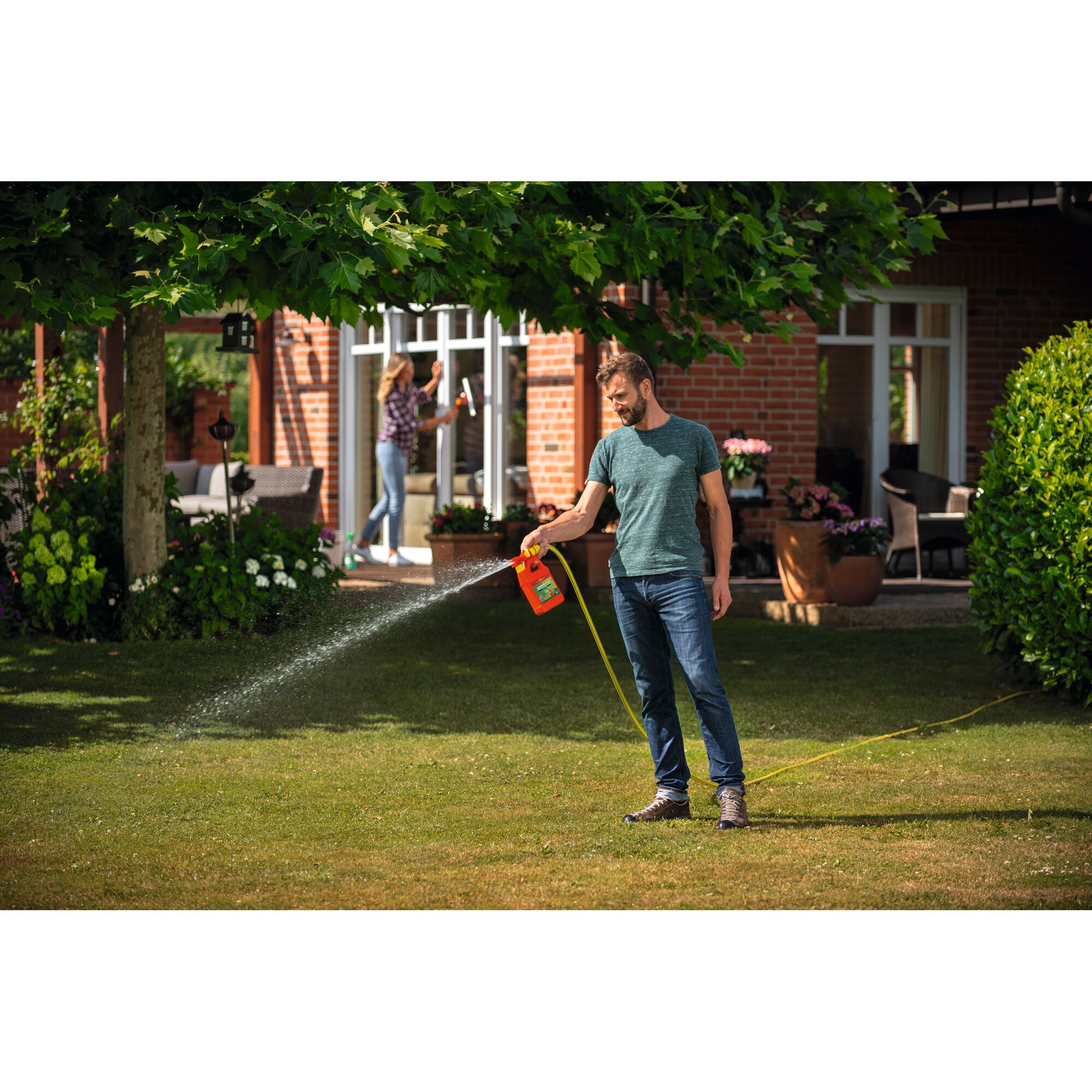 A person is watering the lawn with a WOLFGarten LT100BDA garden sprayer connected to a hose, standing in a garden in front of a brick house with plants and trees. Another person is seen in the background cleaning windows.