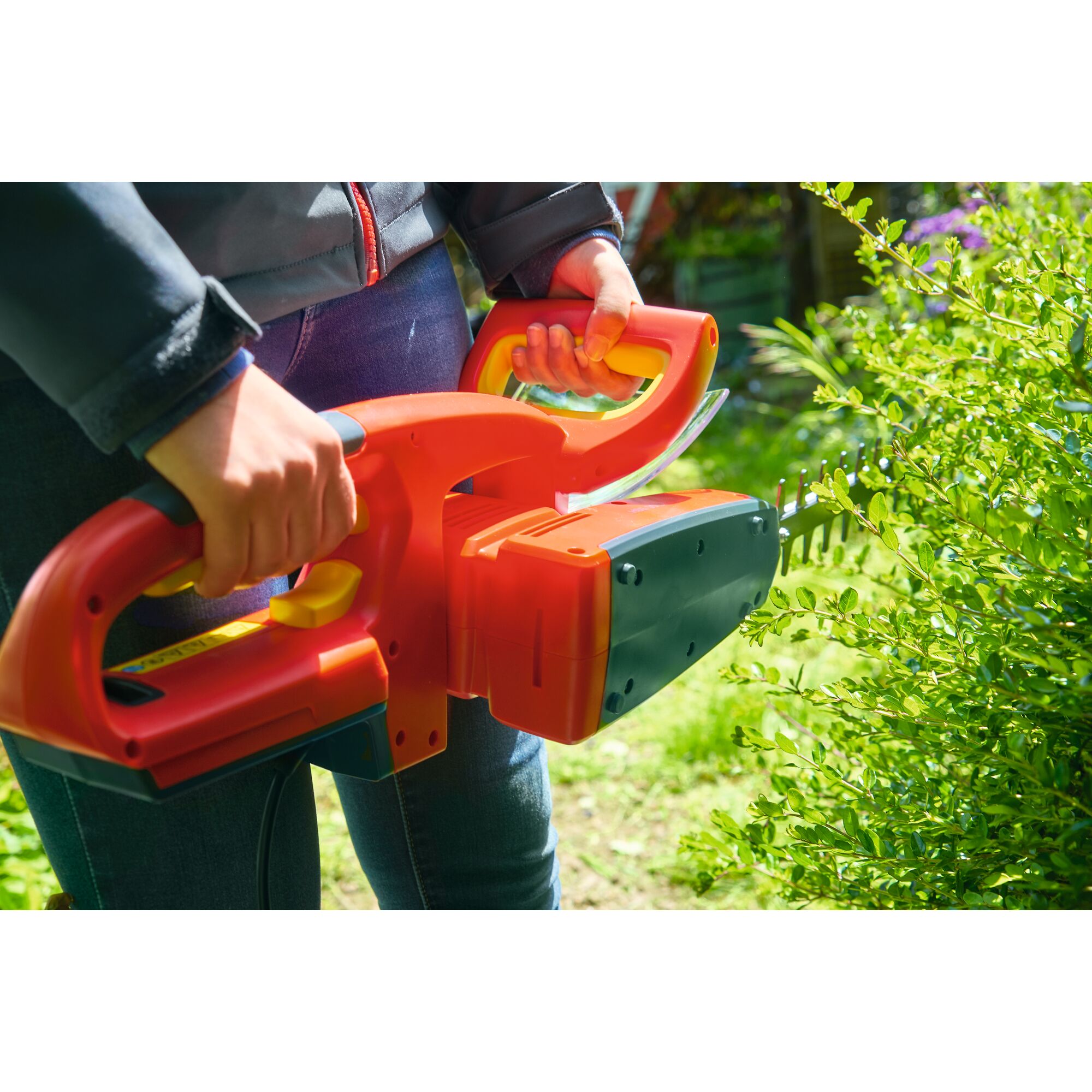 Close-up of a person using an orange and black electric hedge trimmer outdoors to trim green bushes.