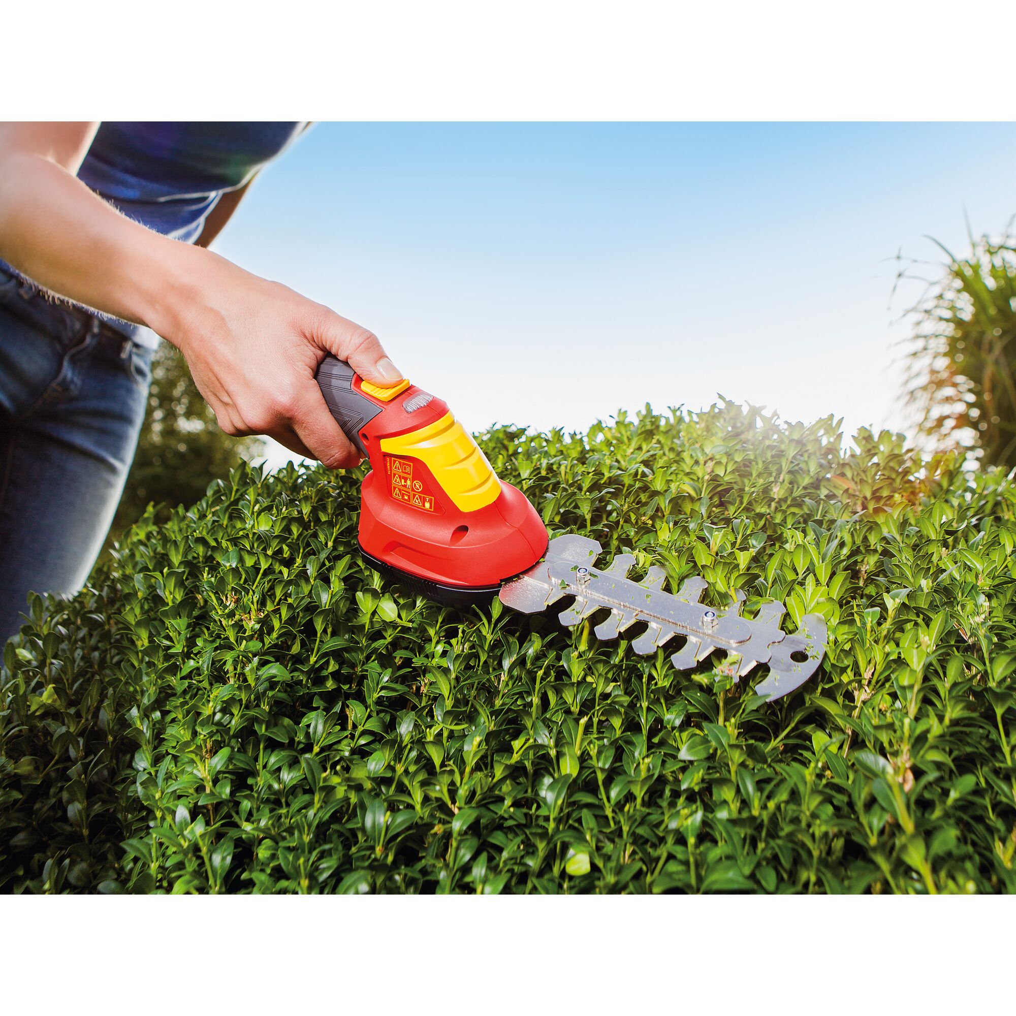 A person uses a WOLFGarten cordless hedge trimmer to trim green bushes outdoors under a bright sky.