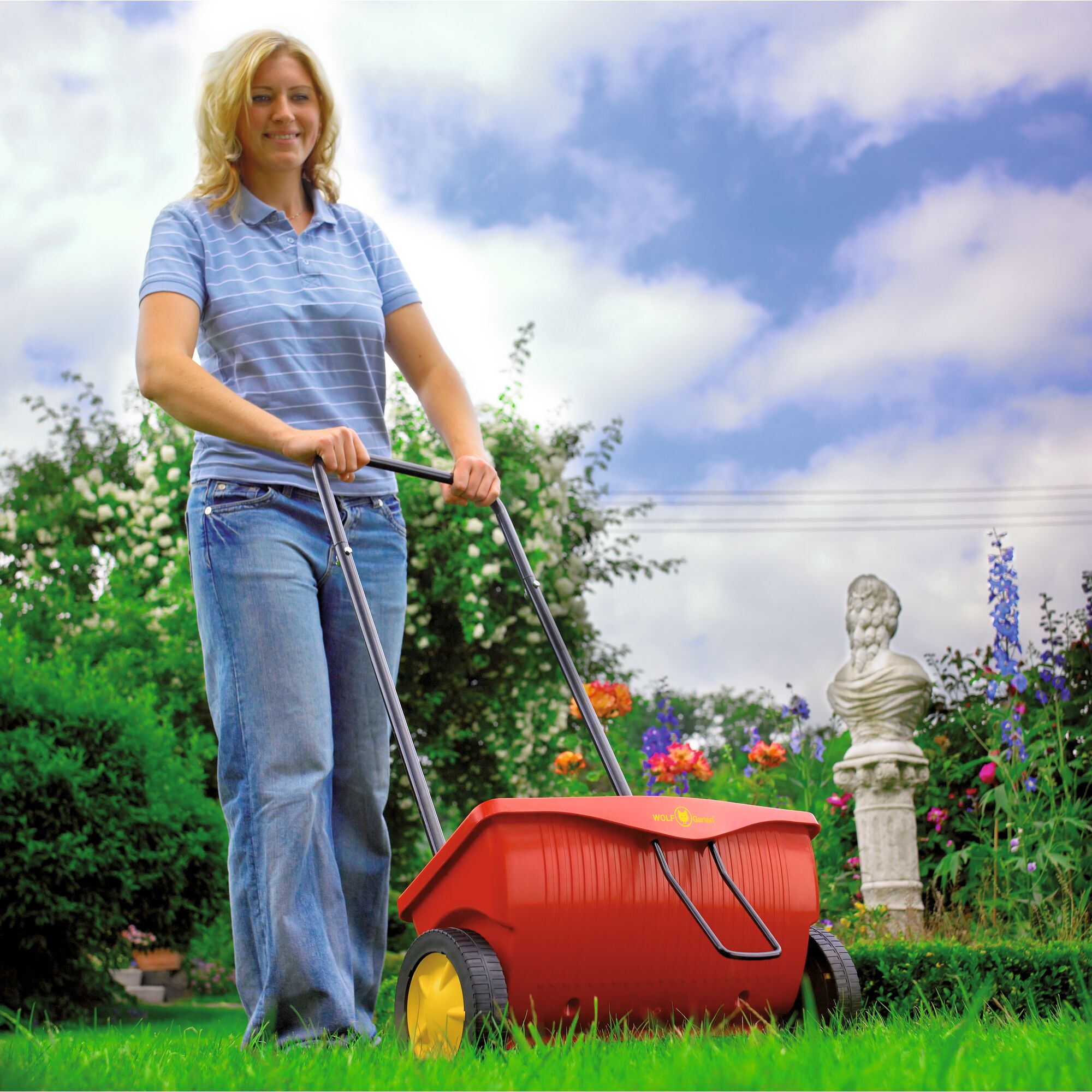 A person is pushing a WOLFGarten WE330 garden spreader with yellow wheels on a lawn in a colorful garden. The main product is the red and yellow garden spreader, used for spreading lawn seed or fertilizer.