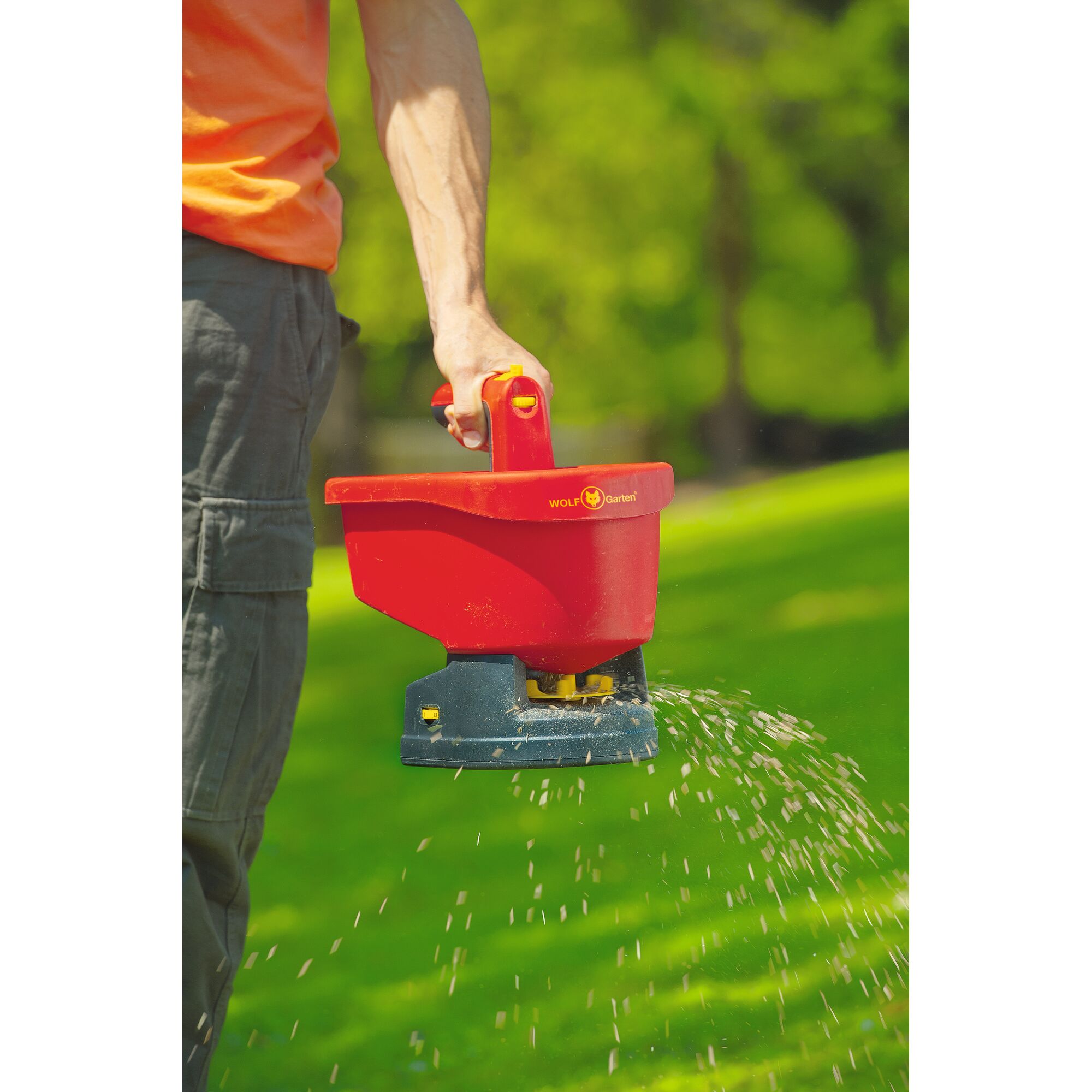 A person is using a WOLFGarten WE-B handheld spreader to distribute seeds or fertilizer outdoors on a grassy area.