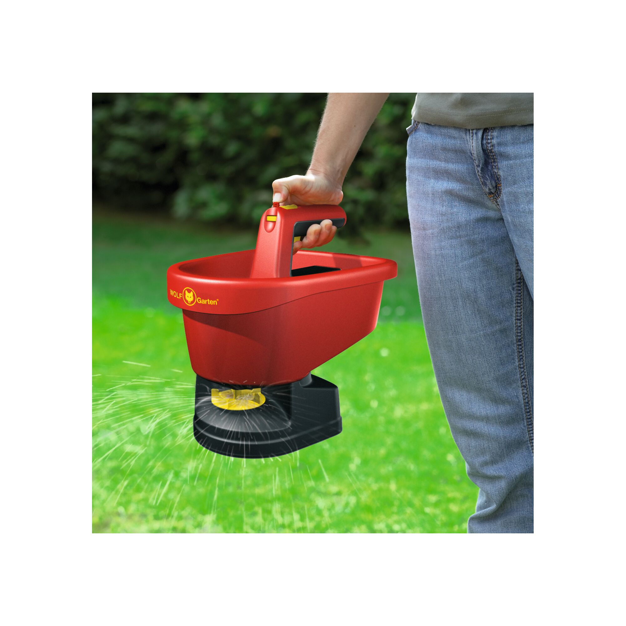A person is holding a red WOLF Garten WE-B handheld spreader, dispensing material onto a grassy lawn outdoors.
