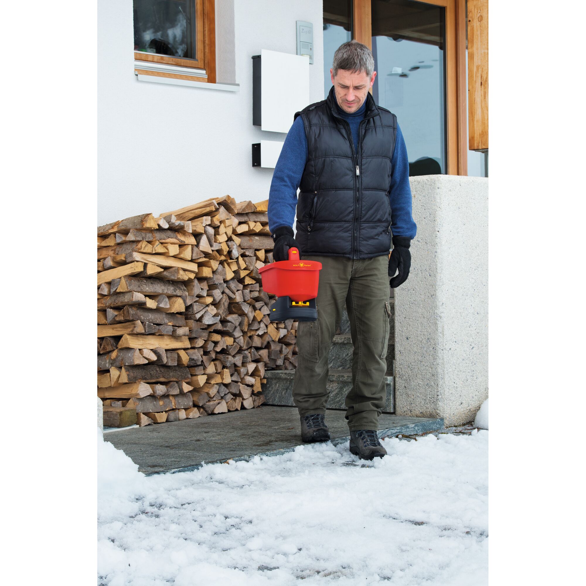 A person standing outdoors on a snowy surface in front of a woodpile, holding a red WOLFGarten WE-B device. The person is wearing a black vest, blue sweater, green pants, gloves, and boots. Their face is obscured.