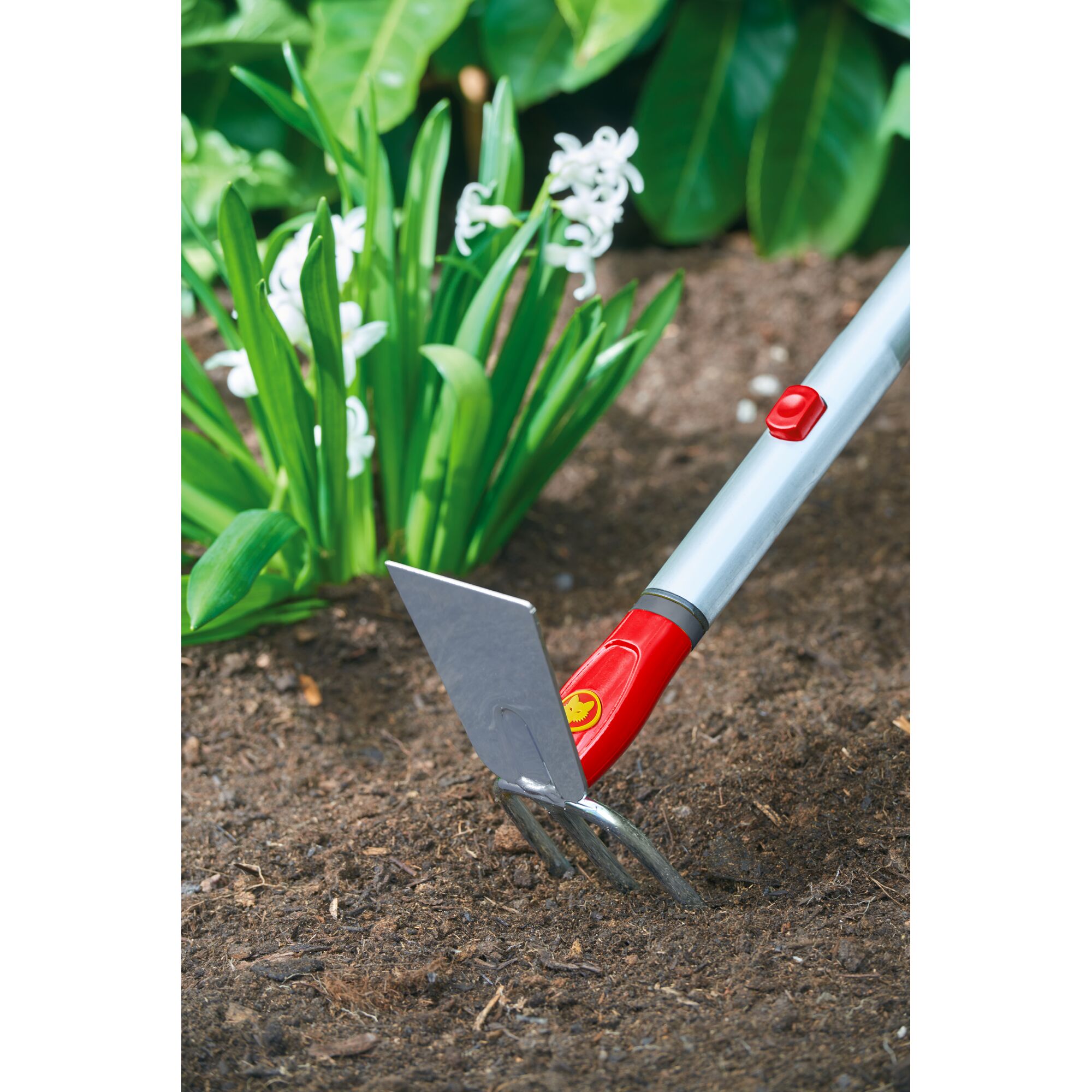 A close-up of a WOLFGarten garden tool with a metal blade and prongs being used in soil near green plants with white flowers.