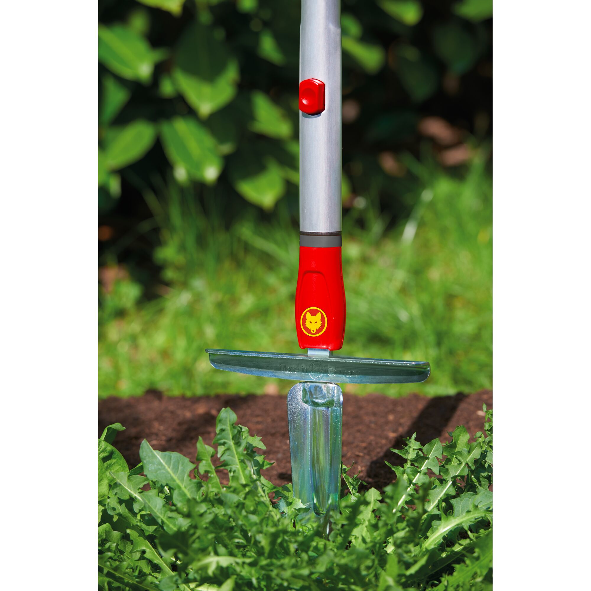 A garden weed puller tool with a metal shaft, red and yellow handle, and metal blade, shown removing weeds from soil in a garden.