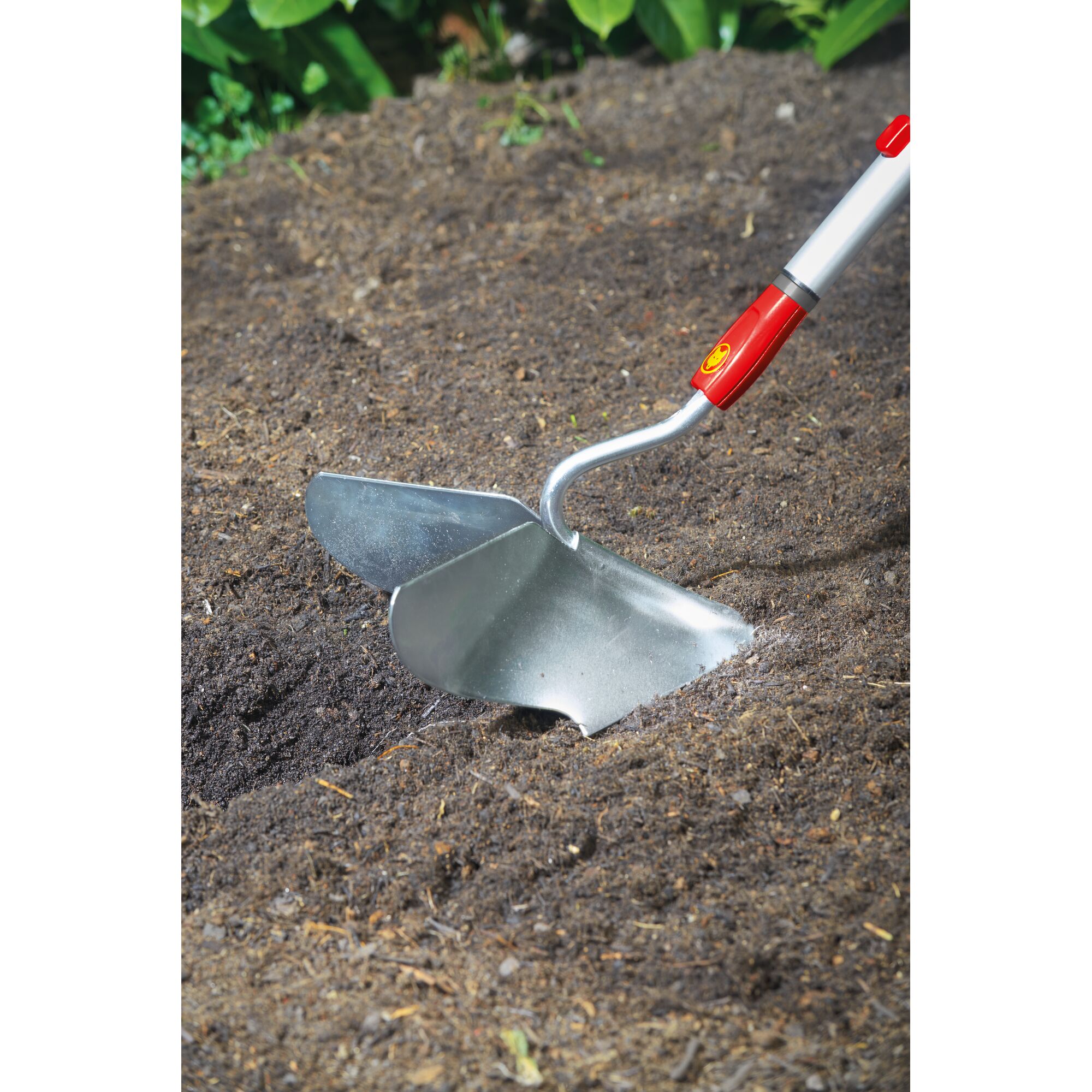 A metal garden hoe with a red and silver handle is being used to dig into soil in a garden bed. The product appears to be from WOLFGarten.