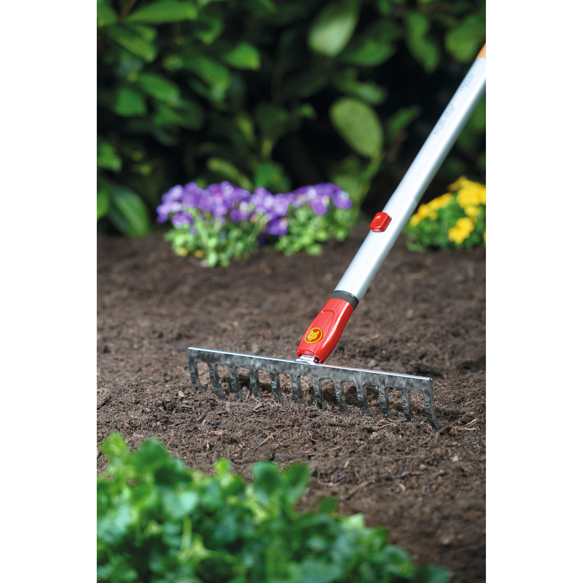 A garden rake with a metal head and a silver handle, featuring red and yellow accents, is being used to level soil in a flower bed with green foliage and blooming flowers in the background.