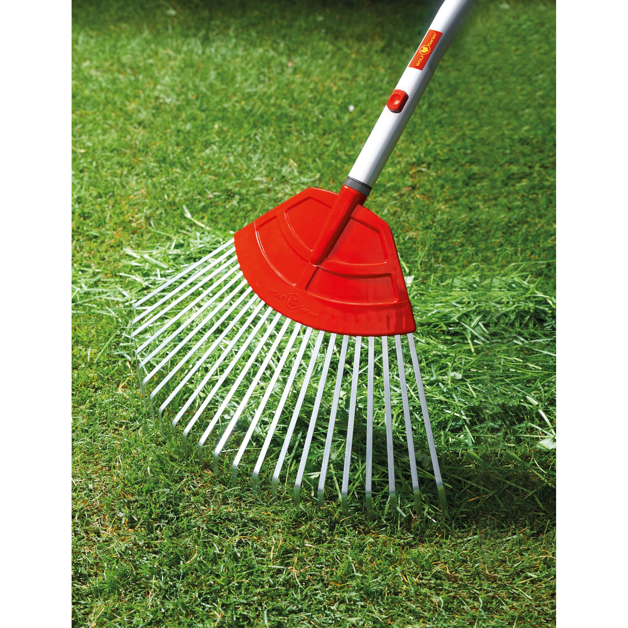 A close-up of a WOLFGarten garden rake with a red plastic head and metal tines, being used on green grass to collect cut grass.